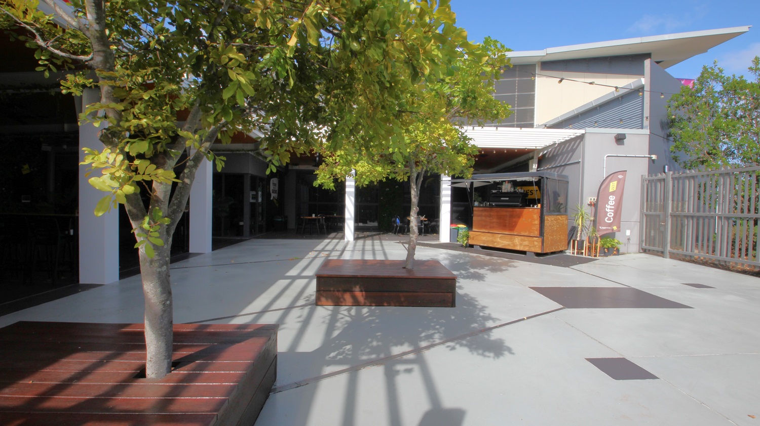 On the left, there are two trees planted in wooden benches. On the right, there is a wooden kiosk with a "Coffee" banner next to it and a grey fence behind. In the middle, there is an open concrete area with shadows of the trees. The background features a building with large windows, a white overhang, and a grey upper section. The floor is light grey concrete, and the ceiling of the kiosk area is white with recessed lights. On the left, there are two trees planted in wooden benches. On the right, there is a wooden kiosk with a "Coffee" banner next to it and a grey fence behind. In the middle, there is an open concrete area with shadows of the trees. The background features a building with large windows, a white overhang, and a grey upper section. The floor is light grey concrete, and the ceiling of the kiosk area is white with recessed lights.