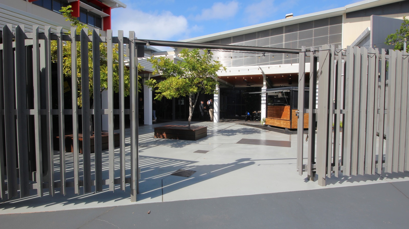 On the left, there is a grey metal fence with vertical slats. On the right, there is a continuation of the same grey metal fence. In the middle, there is an open courtyard with light grey concrete flooring, a wooden bench surrounding a tree, and a person standing near the centre. The background features a building with large windows, a white overhang, and a grey upper section with horizontal slats. On the left, there is a grey metal fence with vertical slats. On the right, there is a continuation of the same grey metal fence. In the middle, there is an open courtyard with light grey concrete flooring, a wooden bench surrounding a tree, and a person standing near the centre. The background features a building with large windows, a white overhang, and a grey upper section with horizontal slats.