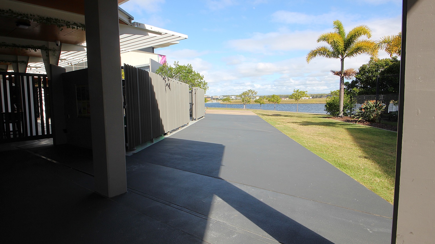 On the left, there is a covered area with a grey metal fence and a white overhang. On the right, there is a grassy lawn with a palm tree and some shrubs. In the middle, there is a wide grey concrete pathway leading towards a body of water in the background. The floor under the covered area is dark grey concrete, and the ceiling is white with recessed lights. On the left, there is a covered area with a grey metal fence and a white overhang. On the right, there is a grassy lawn with a palm tree and some shrubs. In the middle, there is a wide grey concrete pathway leading towards a body of water in the background. The floor under the covered area is dark grey concrete, and the ceiling is white with recessed lights.