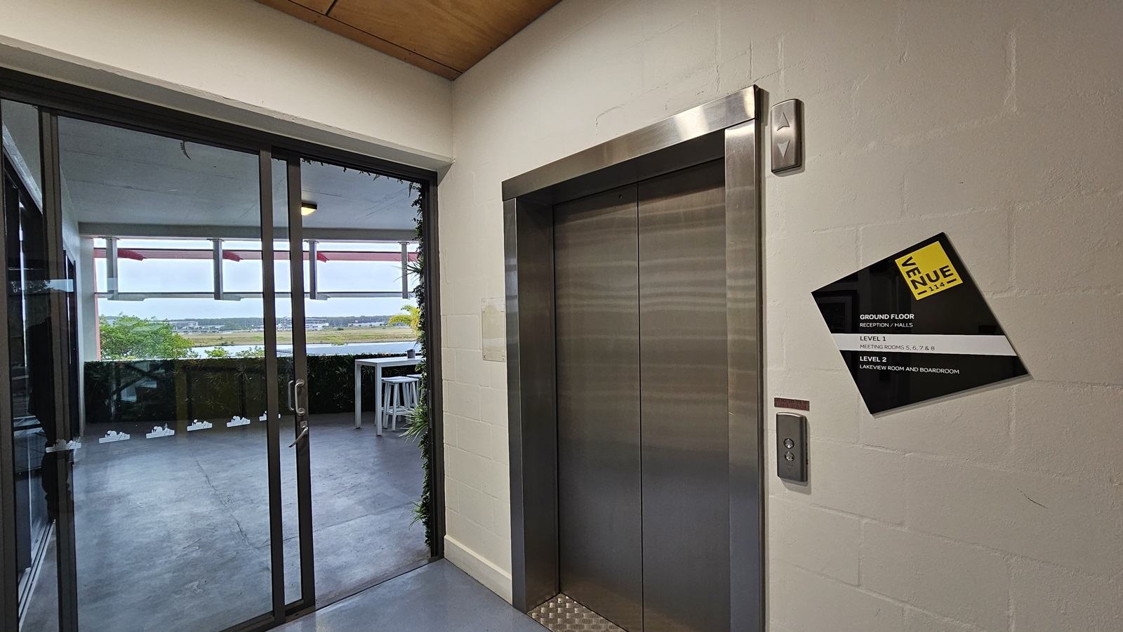 On the left, there is a glass sliding door leading to a balcony with a view of a distant landscape. The balcony has a white table and chairs. On the right, there is a metallic lift with a control panel and a black and yellow sign indicating floor information. In the middle, a white wall separates the lift from the glass door. The floor is grey and appears to be concrete, while the ceiling is wooden. On the left, there is a glass sliding door leading to a balcony with a view of a distant landscape. The balcony has a white table and chairs. On the right, there is a metallic lift with a control panel and a black and yellow sign indicating floor information. In the middle, a white wall separates the lift from the glass door. The floor is grey and appears to be concrete, while the ceiling is wooden.