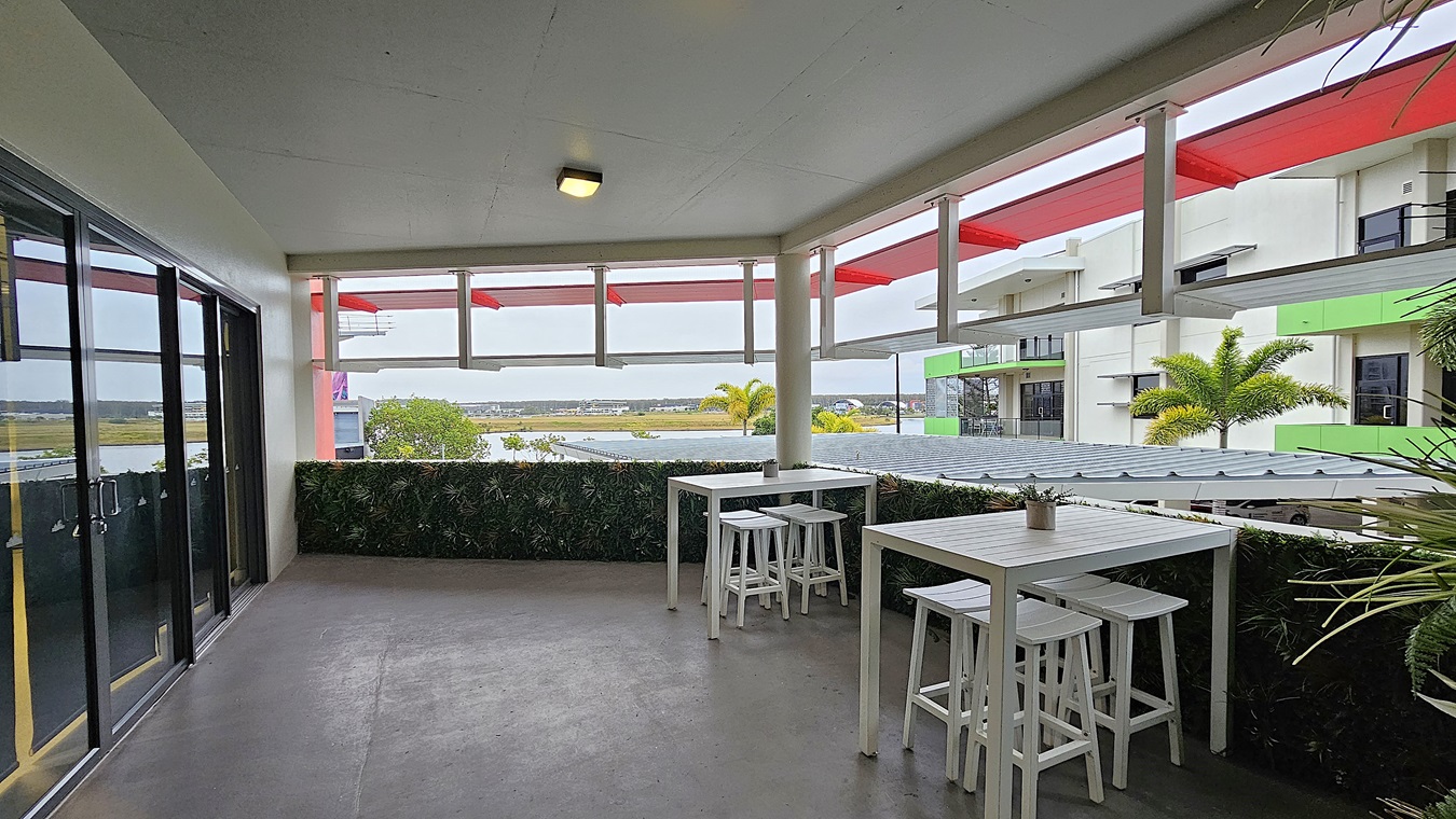 On the left, there are glass sliding doors with black frames. On the right, there are white railings and a view of adjacent buildings with green and white exteriors. In the middle, there are two white high tables with matching stools, and a small plant on one of the tables. The background features a distant landscape with trees and buildings. The floor is grey and appears to be concrete, while the ceiling is white with a single light fixture. On the left, there are glass sliding doors with black frames. On the right, there are white railings and a view of adjacent buildings with green and white exteriors. In the middle, there are two white high tables with matching stools, and a small plant on one of the tables. The background features a distant landscape with trees and buildings. The floor is grey and appears to be concrete, while the ceiling is white with a single light fixture.