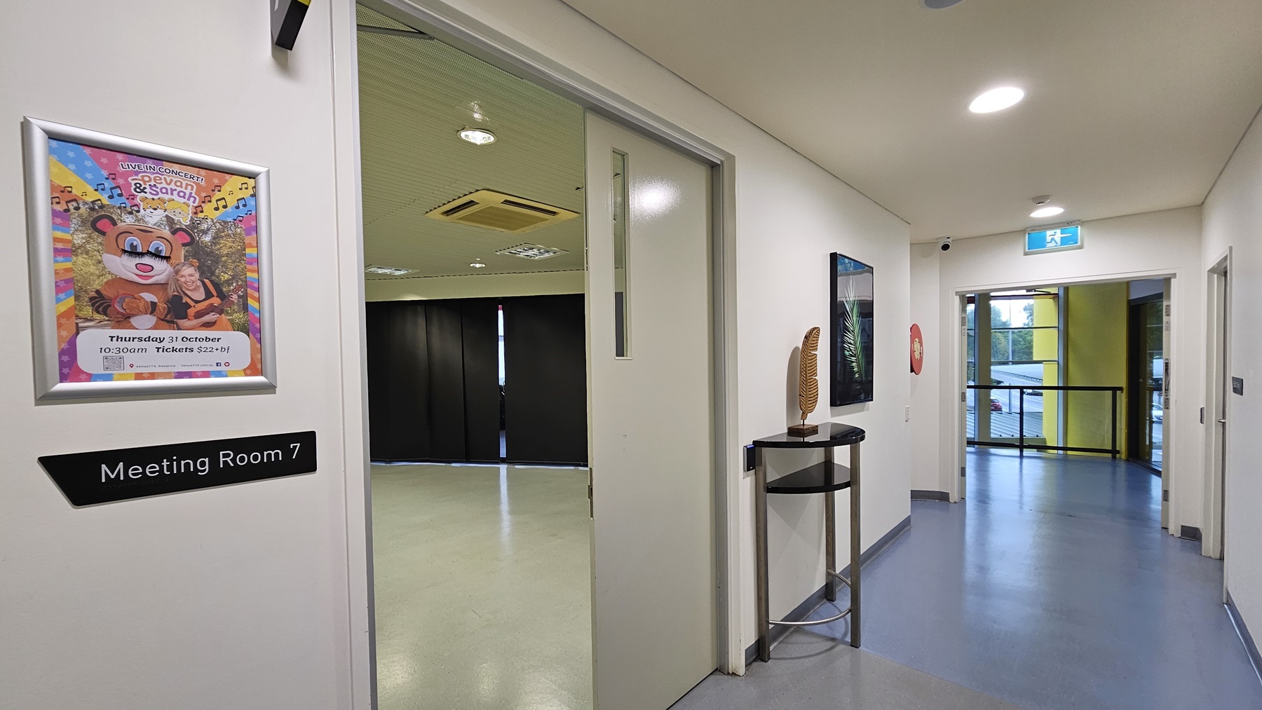 On the left, there is an open door with a sign reading "Meeting Room 7" and a colourful poster above it. Inside the room, there are black curtains covering the windows and a beige floor. On the right, there is a corridor with a fire extinguisher mounted on the wall and a small table with a decorative item. The background features a glass door leading to a balcony with yellow walls. The floor is light grey, and the ceiling is white with recessed lighting and an air conditioning unit. On the left, there is an open door with a sign reading "Meeting Room 7" and a colourful poster above it. Inside the room, there are black curtains covering the windows and a beige floor. On the right, there is a corridor with a fire extinguisher mounted on the wall and a small table with a decorative item. The background features a glass door leading to a balcony with yellow walls. The floor is light grey, and the ceiling is white with recessed lighting and an air conditioning unit.