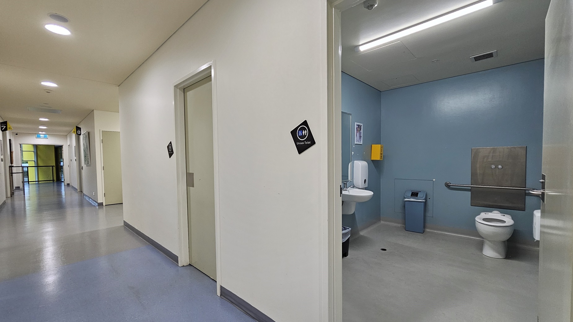 On the left, there is a long corridor with several closed doors, each with a small black sign. The corridor has a light blue and grey floor and white walls. On the right, there is an open door leading to a bathroom with light blue walls, a white toilet, a sink, a soap dispenser, and a blue bin. In the background, the corridor continues with more doors and a bright light at the end. The ceiling is white with recessed lighting and a ventilation unit. On the left, there is a long corridor with several closed doors, each with a small black sign. The corridor has a light blue and grey floor and white walls. On the right, there is an open door leading to a bathroom with light blue walls, a white toilet, a sink, a soap dispenser, and a blue bin. In the background, the corridor continues with more doors and a bright light at the end. The ceiling is white with recessed lighting and a ventilation unit.