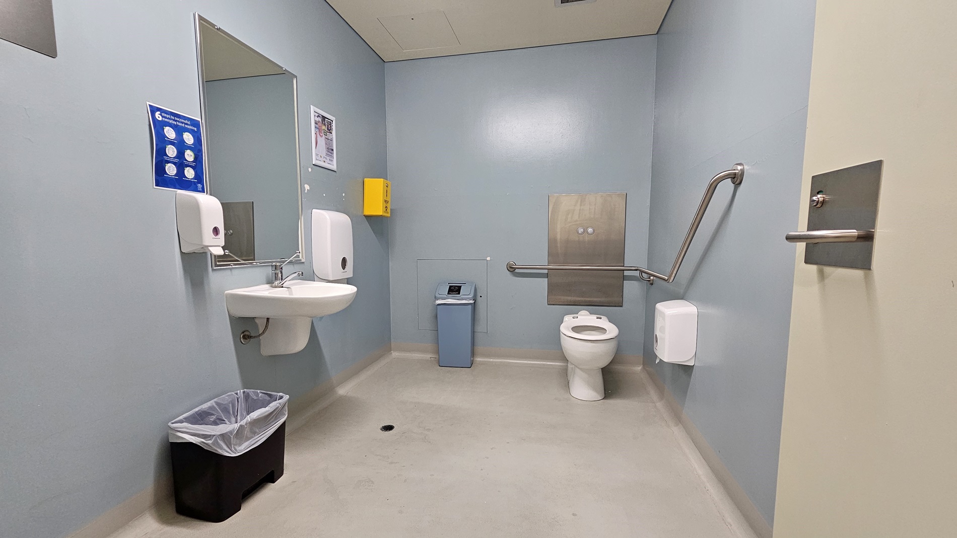 On the left, there is a white sink with a mirror above it, a soap dispenser, and a paper towel dispenser. Below the sink, there is a black bin with a liner. On the right, there is a white toilet with a grab rail on the wall, a toilet paper dispenser, and a small blue bin next to it. In the middle, there is a yellow box mounted on the wall. The floor is light grey, and the ceiling is white with a ventilation panel. On the left, there is a white sink with a mirror above it, a soap dispenser, and a paper towel dispenser. Below the sink, there is a black bin with a liner. On the right, there is a white toilet with a grab rail on the wall, a toilet paper dispenser, and a small blue bin next to it. In the middle, there is a yellow box mounted on the wall. The floor is light grey, and the ceiling is white with a ventilation panel.