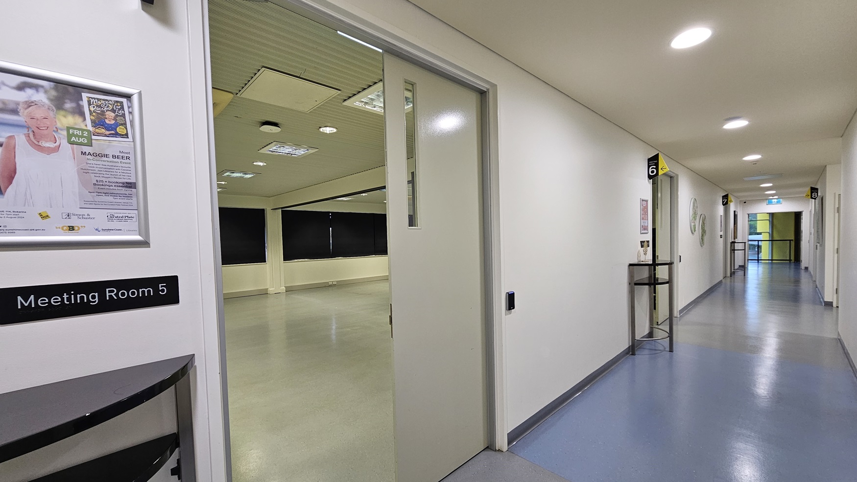 On the left is an open door with a sign reading "Meeting Room 5" and a poster above it. Inside the room, black curtains cover the windows, and a light grey floor. On the right, there is a corridor with a small table holding a decorative item and a sign indicating "6". The corridor has a light blue and grey floor and white walls. In the background, the corridor continues with more doors and a bright light at the end. The ceiling is white with recessed lighting and ventilation panels. On the left is an open door with a sign reading "Meeting Room 5" and a poster above it. Inside the room, black curtains cover the windows, and a light grey floor. On the right, there is a corridor with a small table holding a decorative item and a sign indicating "6". The corridor has a light blue and grey floor and white walls. In the background, the corridor continues with more doors and a bright light at the end. The ceiling is white with recessed lighting and ventilation panels.