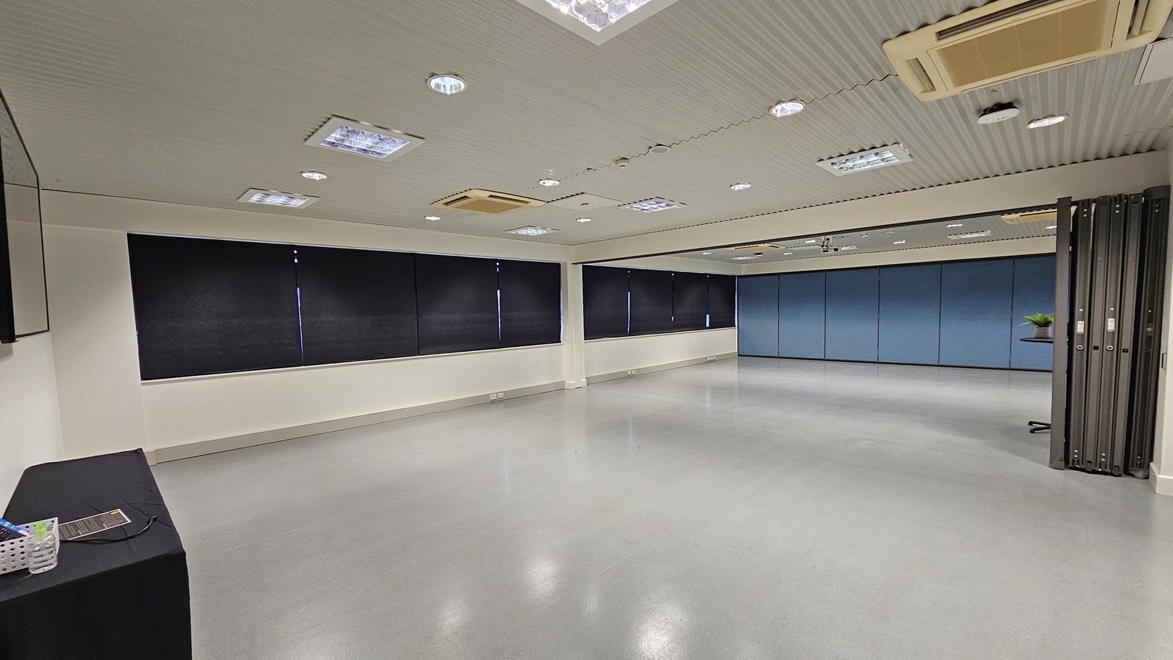 On the left, there is a black table with a basket containing various items. On the right, there is a partition wall with a small table holding a green plant. In the middle, there is a large open space with light grey flooring. The background features black curtains covering windows and a blue partition wall. The ceiling is white with multiple recessed lights and air conditioning units. On the left, there is a black table with a basket containing various items. On the right, there is a partition wall with a small table holding a green plant. In the middle, there is a large open space with light grey flooring. The background features black curtains covering windows and a blue partition wall. The ceiling is white with multiple recessed lights and air conditioning units.