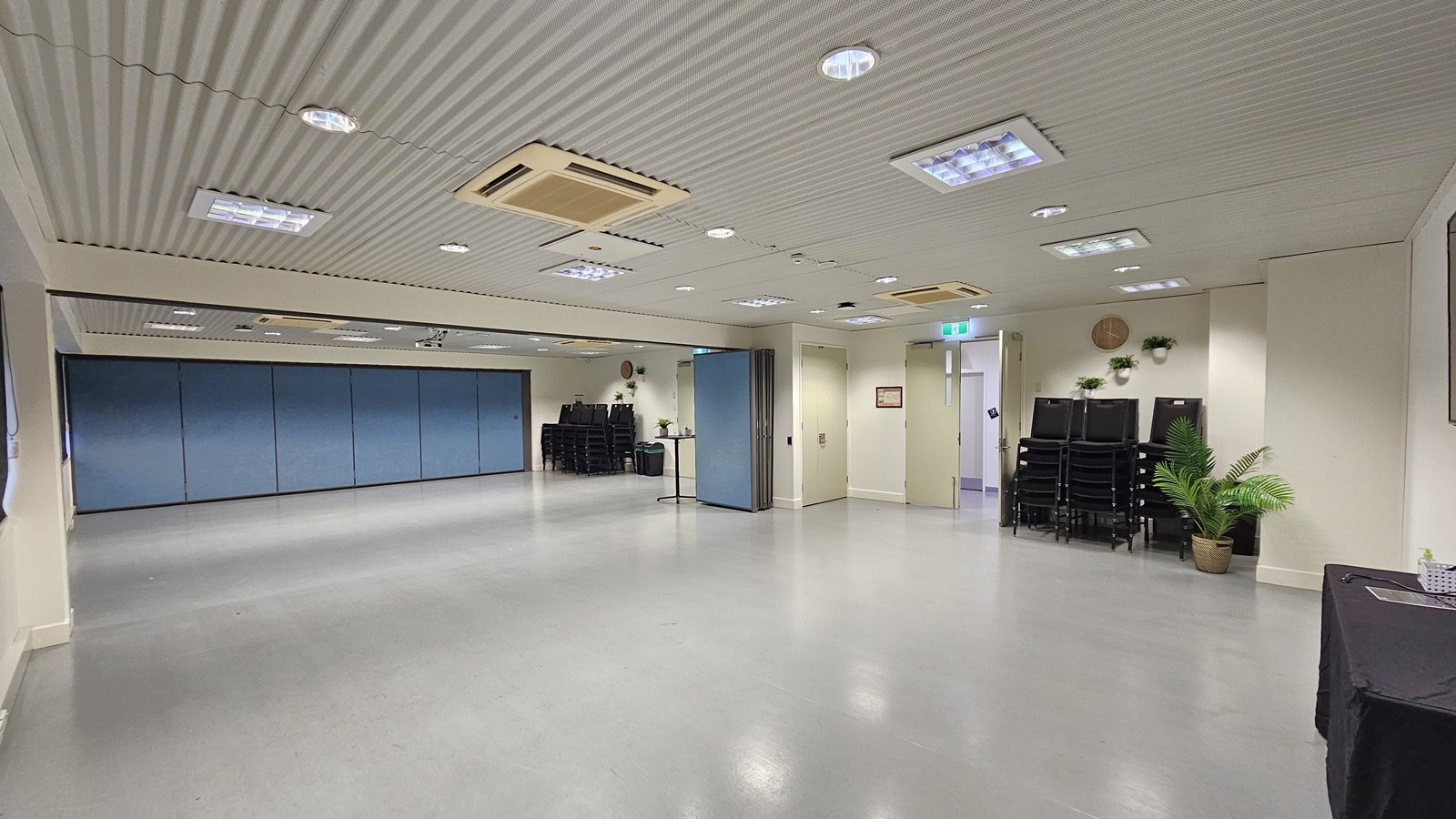 On the left, there is a large open space with blue partition walls. On the right, there is a stack of black chairs next to a potted plant and a small table with a water dispenser. In the middle, there is a column dividing the space. The background features more stacked chairs and a blue partition wall. The floor is light grey, and the ceiling is white with recessed lighting and air conditioning units. On the left, there is a large open space with blue partition walls. On the right, there is a stack of black chairs next to a potted plant and a small table with a water dispenser. In the middle, there is a column dividing the space. The background features more stacked chairs and a blue partition wall. The floor is light grey, and the ceiling is white with recessed lighting and air conditioning units.