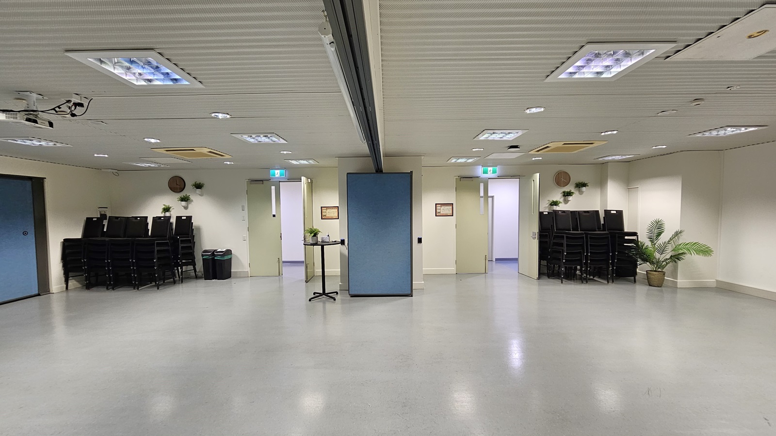 On the left, there is a stack of black chairs next to a black bin and a door with an exit sign above it. On the right, there is another stack of black chairs next to a potted plant and a door with an exit sign above it. In the middle, there is a blue partition wall with a small table holding a plant and a sign on the wall. The floor is light grey, and the ceiling is white with recessed lighting and air conditioning units. On the left, there is a stack of black chairs next to a black bin and a door with an exit sign above it. On the right, there is another stack of black chairs next to a potted plant and a door with an exit sign above it. In the middle, there is a blue partition wall with a small table holding a plant and a sign on the wall. The floor is light grey, and the ceiling is white with recessed lighting and air conditioning units.