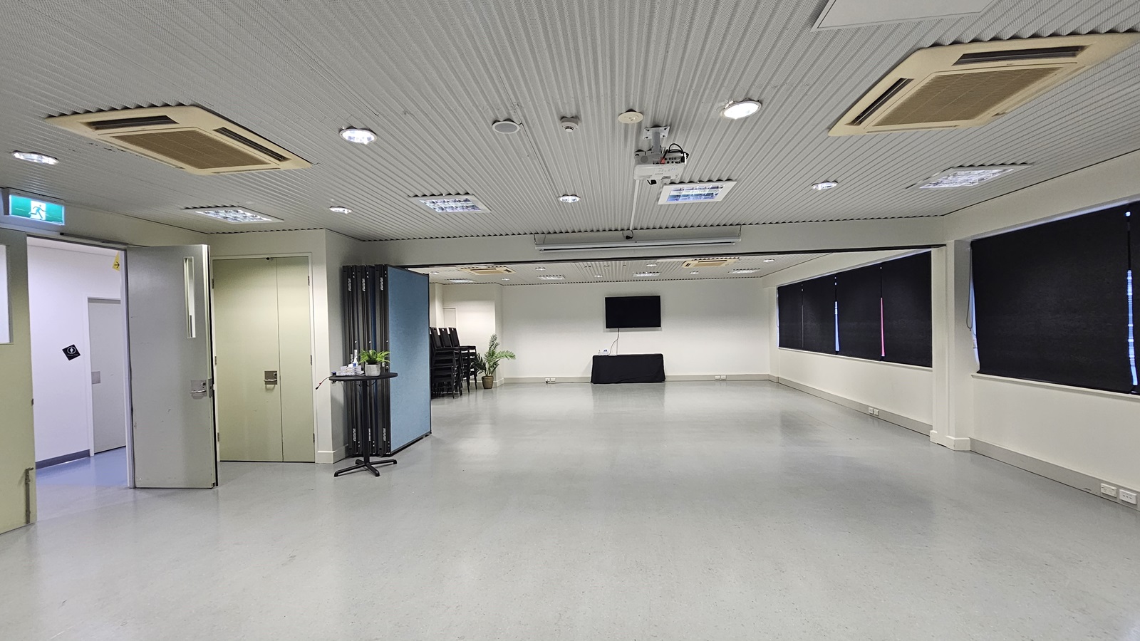On the left, there is a double door with an exit sign above it and a small table with a plant. On the right, there are black curtains covering the windows. In the middle, there is a large open space with a black table against the far wall and a wall-mounted television above it. The background features a stack of black chairs near the back wall. The floor is light grey, and the ceiling is white with recessed lighting and multiple air conditioning units. On the left, there is a double door with an exit sign above it and a small table with a plant. On the right, there are black curtains covering the windows. In the middle, there is a large open space with a black table against the far wall and a wall-mounted television above it. The background features a stack of black chairs near the back wall. The floor is light grey, and the ceiling is white with recessed lighting and multiple air conditioning units.