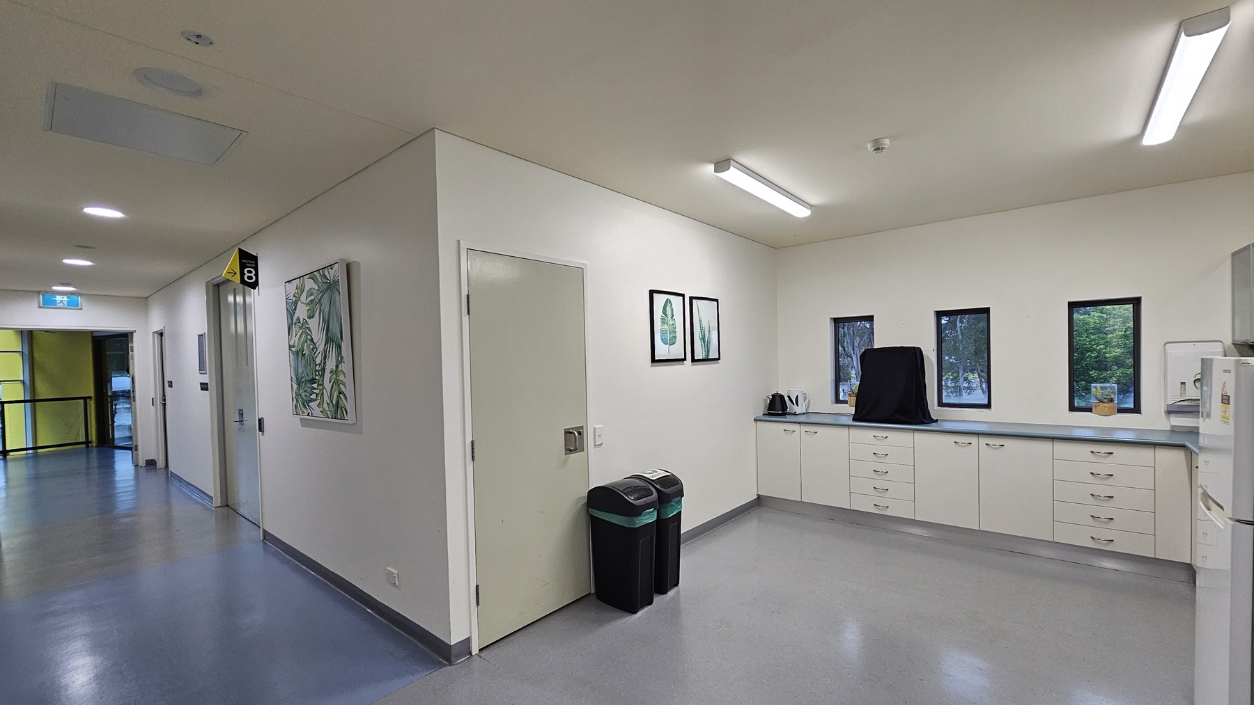 On the left, there is a corridor with white walls, a blue floor, and several doors, one with a sign labelled "8". On the right, there is a kitchenette area with white cabinets, a countertop, a kettle, and three small windows. In the middle, there is a white door next to two black bins with green lids. The background features a yellow door at the end of the corridor. The ceiling is white with recessed lighting and ventilation panels. On the left, there is a corridor with white walls, a blue floor, and several doors, one with a sign labelled "8". On the right, there is a kitchenette area with white cabinets, a countertop, a kettle, and three small windows. In the middle, there is a white door next to two black bins with green lids. The background features a yellow door at the end of the corridor. The ceiling is white with recessed lighting and ventilation panels.
