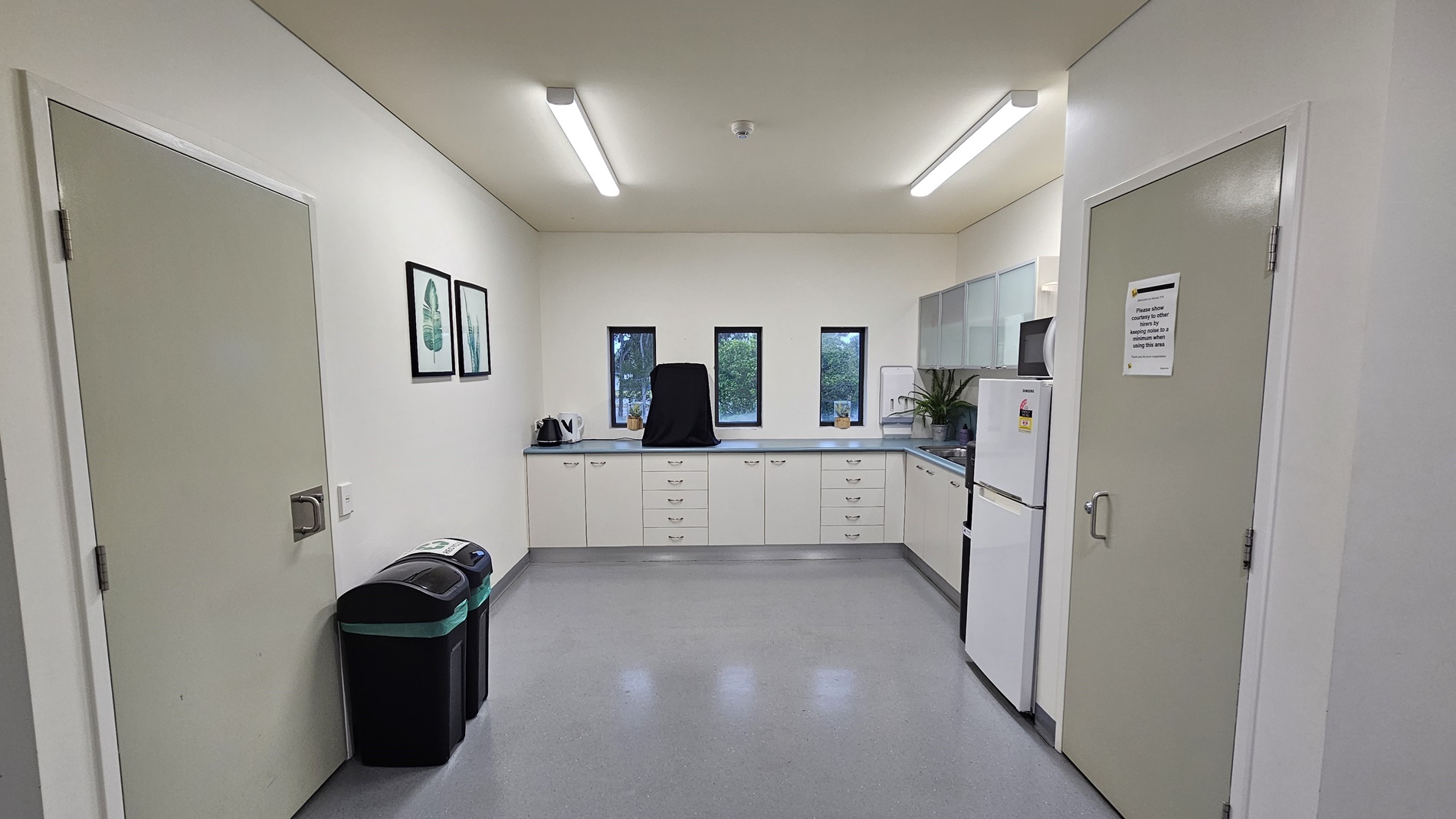 On the left, there is a white door next to two black bins with green lids. On the right, there is another white door with a sign on it and a white refrigerator beside it. In the middle, there is a kitchenette with white cabinets, a countertop, a kettle, and three small windows above the counter. The floor is light grey, and the ceiling is white with recessed lighting. On the left, there is a white door next to two black bins with green lids. On the right, there is another white door with a sign on it and a white refrigerator beside it. In the middle, there is a kitchenette with white cabinets, a countertop, a kettle, and three small windows above the counter. The floor is light grey, and the ceiling is white with recessed lighting.