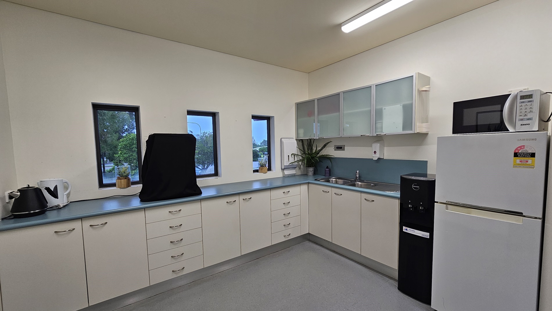 On the left, there is a white kettle and a small potted plant on a blue countertop. On the right, there is a white refrigerator with a microwave on top and a black water dispenser beside it. In the middle, there are three small windows above the countertop, a sink with a plant next to it, and white cabinets with frosted glass doors. The floor is light grey, and the ceiling is white with a long fluorescent light fixture. On the left, there is a white kettle and a small potted plant on a blue countertop. On the right, there is a white refrigerator with a microwave on top and a black water dispenser beside it. In the middle, there are three small windows above the countertop, a sink with a plant next to it, and white cabinets with frosted glass doors. The floor is light grey, and the ceiling is white with a long fluorescent light fixture.