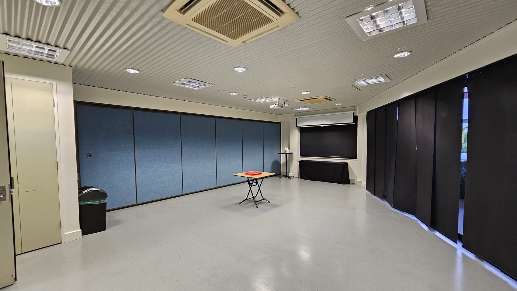 On the left, there is a white door next to a black bin with a green lid. On the right, there are black curtains covering the windows. In the middle, there is a small table with a red object on it. The background features blue partition walls and a black table with a whiteboard above it. The floor is light grey, and the ceiling is white with recessed lighting and an air conditioning unit. On the left, there is a white door next to a black bin with a green lid. On the right, there are black curtains covering the windows. In the middle, there is a small table with a red object on it. The background features blue partition walls and a black table with a whiteboard above it. The floor is light grey, and the ceiling is white with recessed lighting and an air conditioning unit.