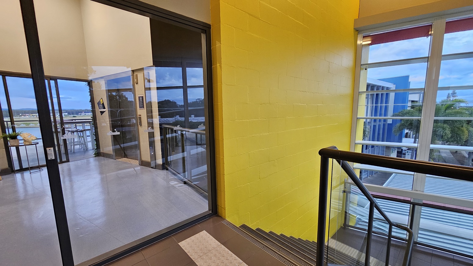 On the left, there is a glass door leading to a room with grey flooring, white tables, and chairs. On the right, there is a yellow brick wall next to a staircase with metal railings. In the background, large windows reveal a view of blue buildings and palm trees. The floor in the foreground is dark grey, and the ceiling is white with recessed lighting. On the left, there is a glass door leading to a room with grey flooring, white tables, and chairs. On the right, there is a yellow brick wall next to a staircase with metal railings. In the background, large windows reveal a view of blue buildings and palm trees. The floor in the foreground is dark grey, and the ceiling is white with recessed lighting.