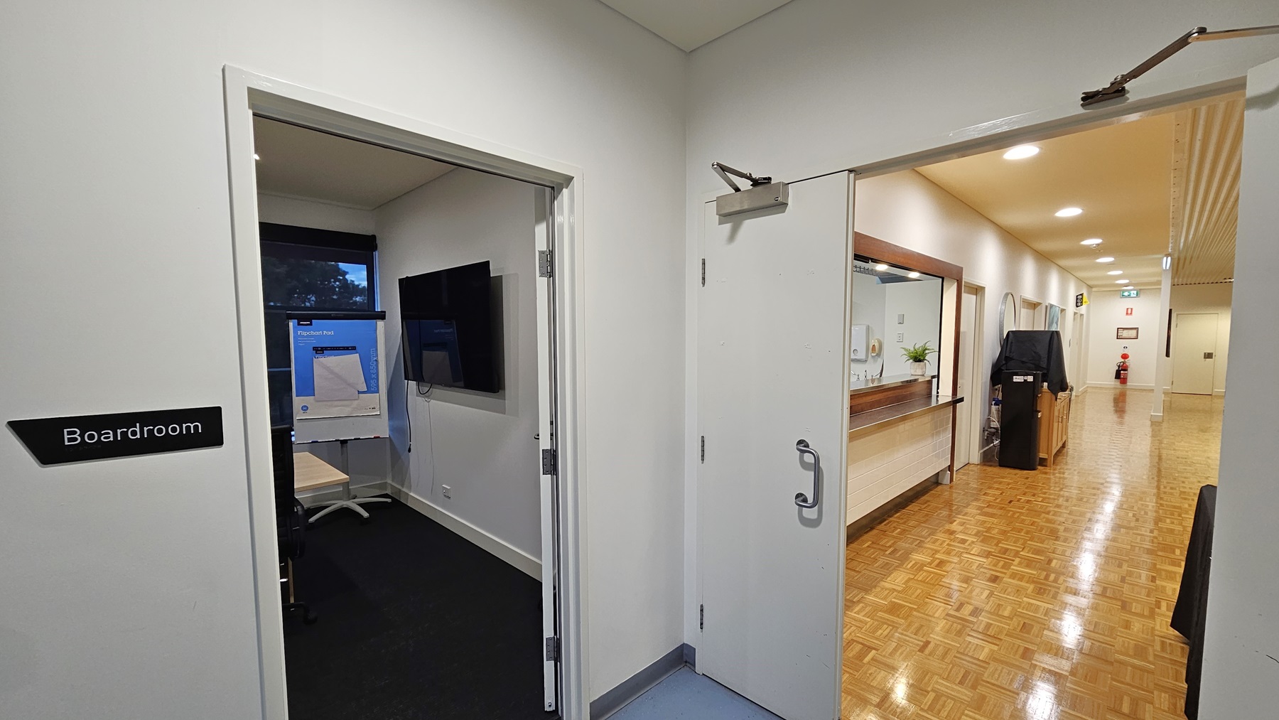 On the left, there is an open door with a sign labelled "Boardroom," revealing a room with a black carpet, a wall-mounted television, and a flip chart. On the right, there is a corridor with a wooden parquet floor, white walls, and a reception desk with a mirror above it. In the background, the corridor extends to a fire extinguisher and an exit sign. The ceiling is white with recessed lighting. On the left, there is an open door with a sign labelled "Boardroom," revealing a room with a black carpet, a wall-mounted television, and a flip chart. On the right, there is a corridor with a wooden parquet floor, white walls, and a reception desk with a mirror above it. In the background, the corridor extends to a fire extinguisher and an exit sign. The ceiling is white with recessed lighting.