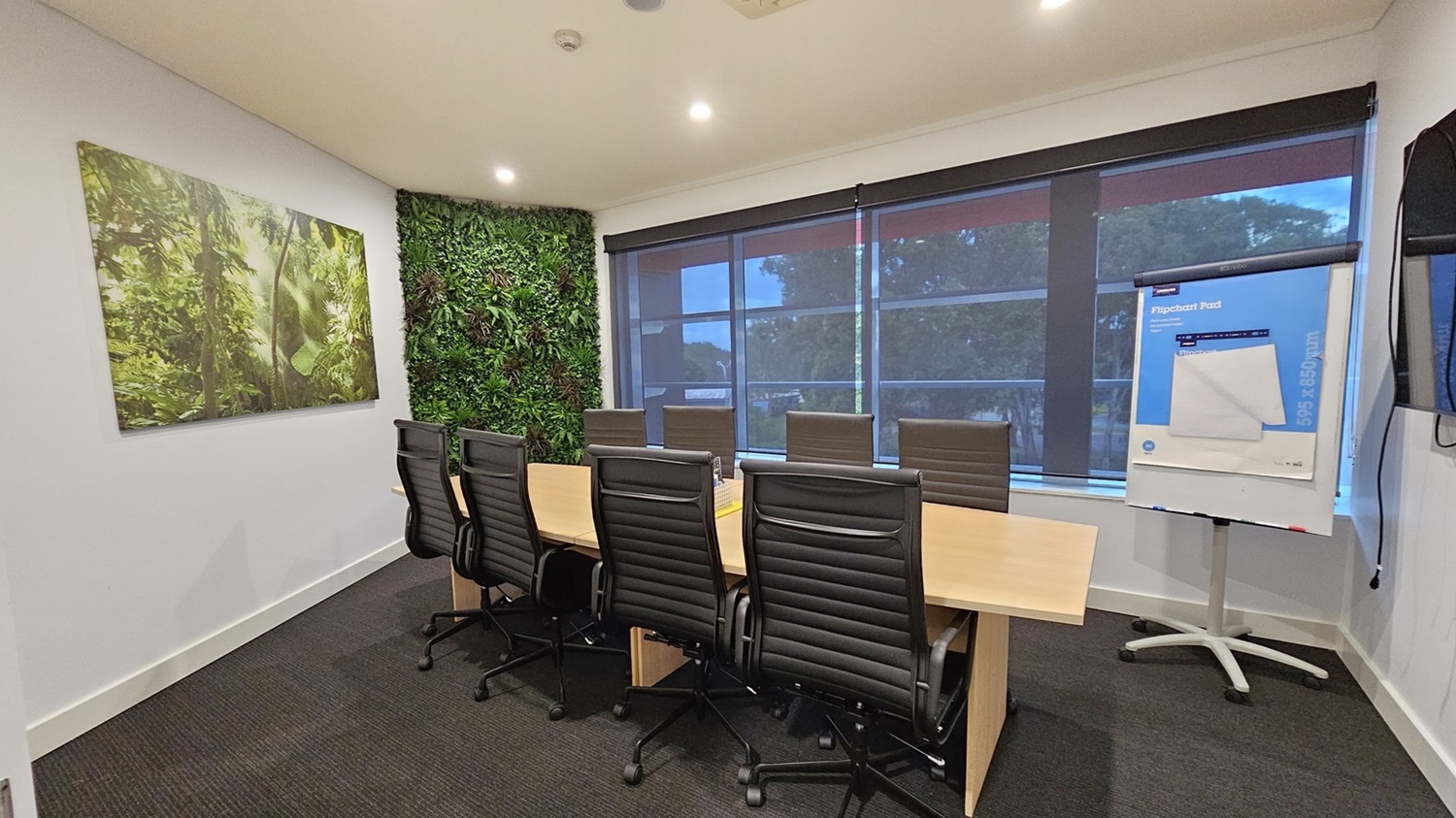 On the left, there is a large framed picture of a forest scene on a white wall and a section of wall covered in green foliage. On the right, there are large windows with roller blinds partially down and a flip chart on a stand. In the middle, there is a long wooden conference table surrounded by black office chairs. The floor is covered in dark grey carpet, and the ceiling is white with recessed lighting. On the left, there is a large framed picture of a forest scene on a white wall and a section of wall covered in green foliage. On the right, there are large windows with roller blinds partially down and a flip chart on a stand. In the middle, there is a long wooden conference table surrounded by black office chairs. The floor is covered in dark grey carpet, and the ceiling is white with recessed lighting.