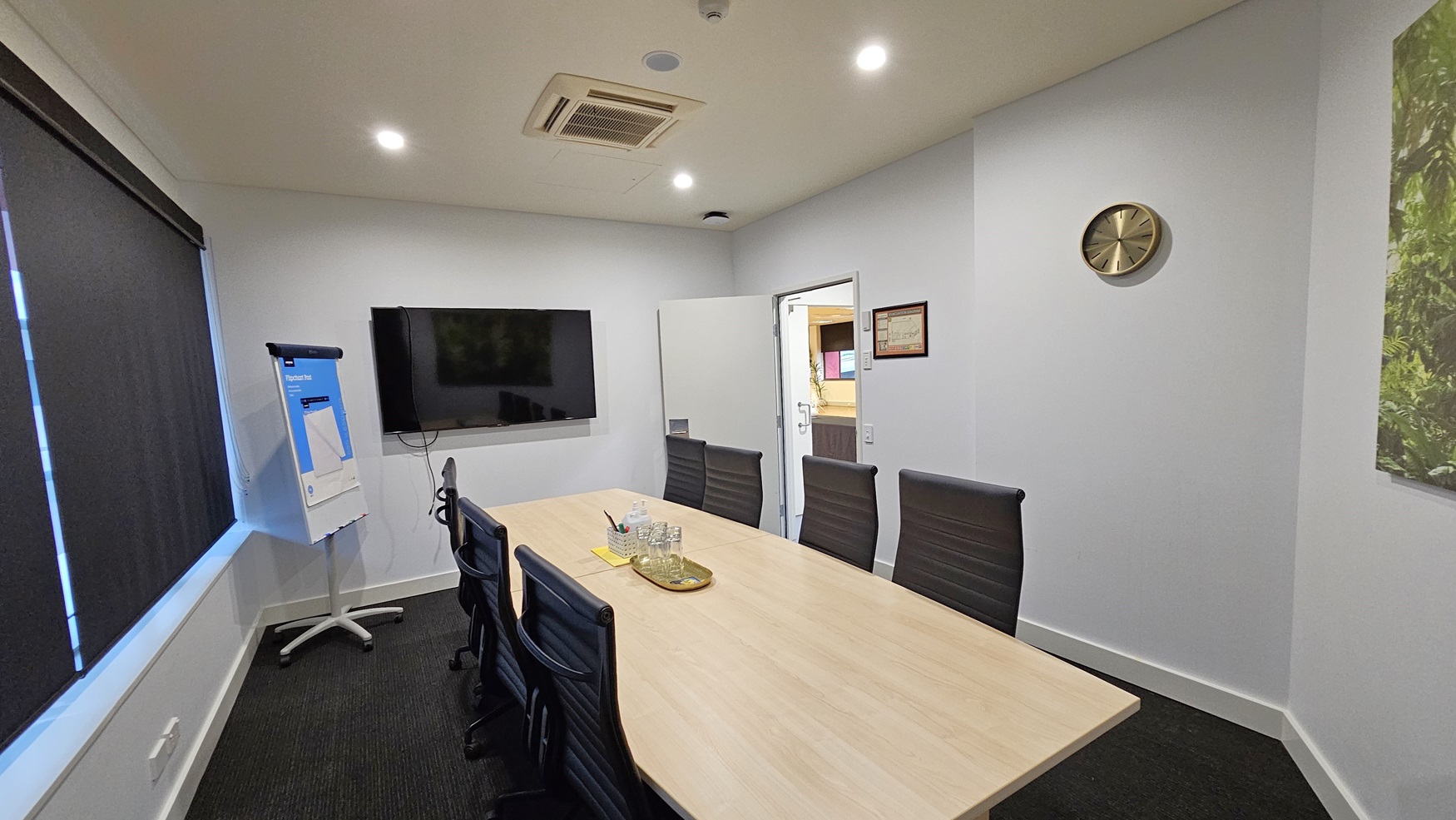 On the left, there is a flip chart on a stand next to a large wall-mounted television. On the right, there is a clock on the wall and a framed certificate above an open door leading to another room. In the middle, there is a long wooden conference table with black office chairs around it, and a tray with glasses and a water jug on the table. The floor is covered in dark grey carpet, and the ceiling is white with recessed lighting and an air conditioning unit. On the left, there is a flip chart on a stand next to a large wall-mounted television. On the right, there is a clock on the wall and a framed certificate above an open door leading to another room. In the middle, there is a long wooden conference table with black office chairs around it, and a tray with glasses and a water jug on the table. The floor is covered in dark grey carpet, and the ceiling is white with recessed lighting and an air conditioning unit.