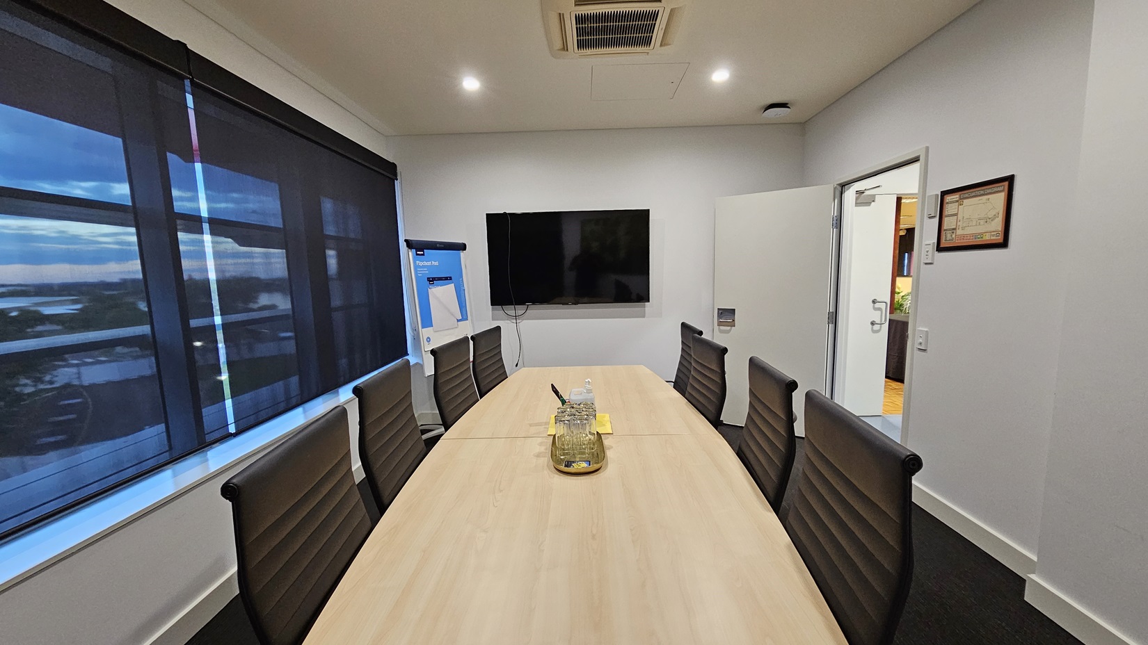 On the left, there are large windows with black roller blinds partially down and a flip chart on a stand. On the right, there is an open door leading to another room and a framed certificate on the wall. In the middle, there is a long wooden conference table with black office chairs around it, and a tray with a water jug and glasses on the table. The floor is covered in dark grey carpet, and the ceiling is white with recessed lighting and an air conditioning unit. On the left, there are large windows with black roller blinds partially down and a flip chart on a stand. On the right, there is an open door leading to another room and a framed certificate on the wall. In the middle, there is a long wooden conference table with black office chairs around it, and a tray with a water jug and glasses on the table. The floor is covered in dark grey carpet, and the ceiling is white with recessed lighting and an air conditioning unit.