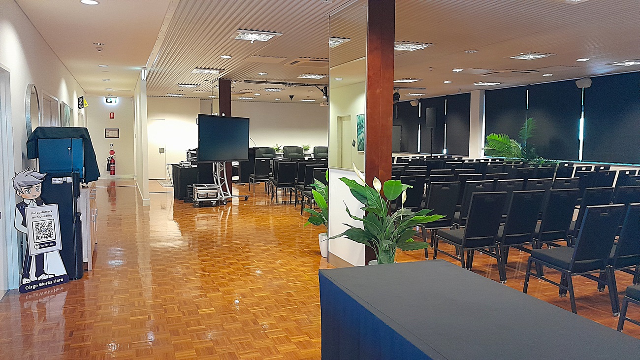 On the left, there is a corridor with a wooden parquet floor, white walls, and a blue recycling bin with a cartoon character on it. On the right, there are rows of black chairs facing a large screen, with black roller blinds partially down on the windows. In the middle, there is a large television on a stand and a potted plant near a wooden pillar. The ceiling is white with recessed lighting and ventilation panels. On the left, there is a corridor with a wooden parquet floor, white walls, and a blue recycling bin with a cartoon character on it. On the right, there are rows of black chairs facing a large screen, with black roller blinds partially down on the windows. In the middle, there is a large television on a stand and a potted plant near a wooden pillar. The ceiling is white with recessed lighting and ventilation panels.