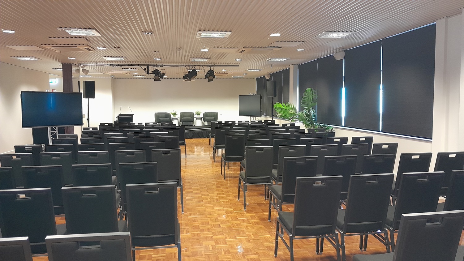 On the left, there is a large television on a stand and a black speaker. On the right, there are large windows with black roller blinds partially down and a potted plant near the corner. In the middle, there are rows of black chairs facing a stage with a podium, three black chairs, and a small table. The floor is covered in wooden parquet, and the ceiling is white with recessed lighting and ventilation panels. On the left, there is a large television on a stand and a black speaker. On the right, there are large windows with black roller blinds partially down and a potted plant near the corner. In the middle, there are rows of black chairs facing a stage with a podium, three black chairs, and a small table. The floor is covered in wooden parquet, and the ceiling is white with recessed lighting and ventilation panels.