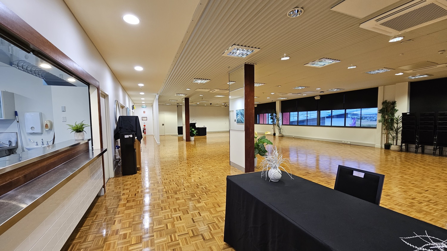 On the left, there is a long counter with a service window, a potted plant, and a black cabinet. On the right, there are large windows with black roller blinds partially down and a stack of black chairs in the corner. In the middle, there is a black table with a decorative vase and a single black chair. The floor is covered in wooden parquet, and the ceiling is white with recessed lighting and ventilation panels. On the left, there is a long counter with a service window, a potted plant, and a black cabinet. On the right, there are large windows with black roller blinds partially down and a stack of black chairs in the corner. In the middle, there is a black table with a decorative vase and a single black chair. The floor is covered in wooden parquet, and the ceiling is white with recessed lighting and ventilation panels.