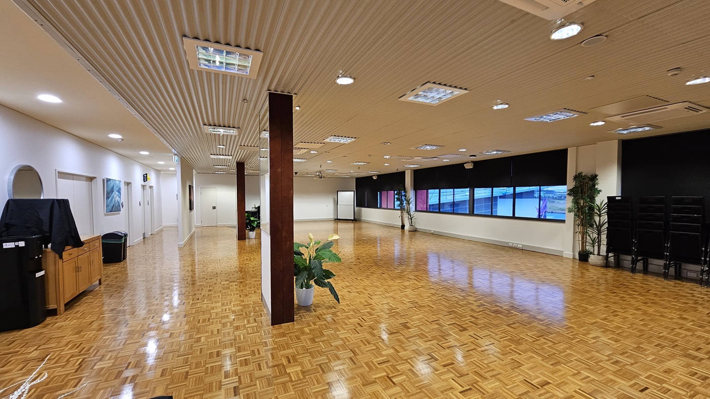 On the left, there is a wooden cabinet with a black cover, a potted plant, and a black bin. On the right, there are large windows with black roller blinds partially down, a stack of black chairs, and two potted plants. In the middle, there is a wooden pillar with a potted plant at its base. The floor is covered in wooden parquet, and the ceiling is white with recessed lighting and ventilation panels. On the left, there is a wooden cabinet with a black cover, a potted plant, and a black bin. On the right, there are large windows with black roller blinds partially down, a stack of black chairs, and two potted plants. In the middle, there is a wooden pillar with a potted plant at its base. The floor is covered in wooden parquet, and the ceiling is white with recessed lighting and ventilation panels.