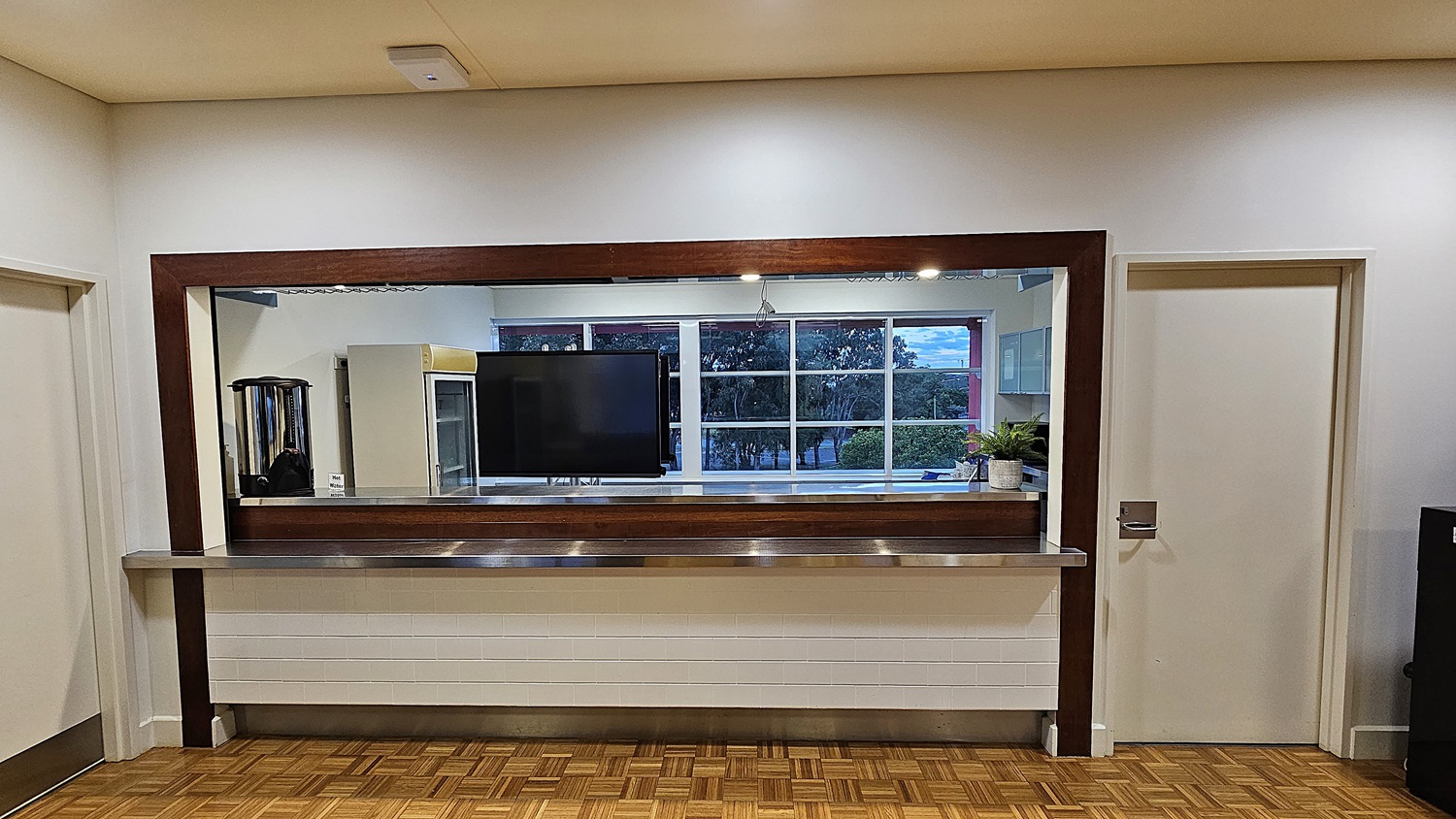 On the left, there is a white door with a silver handle. On the right, there is another white door with a silver handle and a black cabinet next to it. In the middle, there is a large rectangular mirror with a wooden frame above a long stainless steel counter. Behind the mirror, there is a television mounted on the wall and a potted plant on a shelf. The floor is covered in wooden parquet, and the ceiling is beige with recessed lighting. On the left, there is a white door with a silver handle. On the right, there is another white door with a silver handle and a black cabinet next to it. In the middle, there is a large rectangular mirror with a wooden frame above a long stainless steel counter. Behind the mirror, there is a television mounted on the wall and a potted plant on a shelf. The floor is covered in wooden parquet, and the ceiling is beige with recessed lighting.