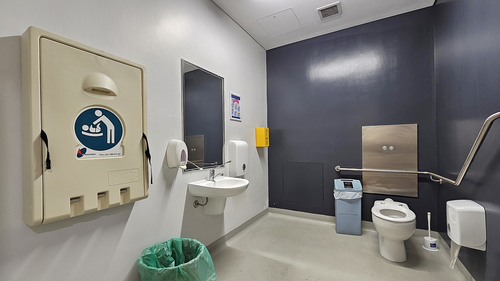 On the left, there is a beige baby changing station mounted on the wall, a green bin on the floor, a sink with a mirror above it, and a soap dispenser. On the right, there is a white toilet with a grey grab rail next to it, a blue bin, and a toilet paper dispenser. In the middle, there is a yellow wall-mounted box. The floor is light grey, and the ceiling is white with recessed lighting and a ventilation panel. On the left, there is a beige baby changing station mounted on the wall, a green bin on the floor, a sink with a mirror above it, and a soap dispenser. On the right, there is a white toilet with a grey grab rail next to it, a blue bin, and a toilet paper dispenser. In the middle, there is a yellow wall-mounted box. The floor is light grey, and the ceiling is white with recessed lighting and a ventilation panel.