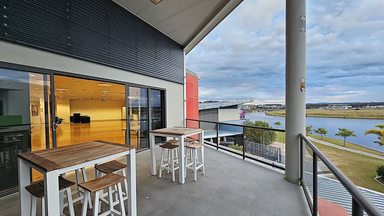 On the left, there is a glass door leading to an indoor area with a wooden floor and ceiling lights. On the right, there is a view of a balcony with a metal railing, overlooking a grassy area with trees and a body of water. In the foreground, there are two white high tables with wooden tops and matching stools. The floor of the balcony is grey concrete, and the ceiling above the balcony is white with recessed lighting. On the left, there is a glass door leading to an indoor area with a wooden floor and ceiling lights. On the right, there is a view of a balcony with a metal railing, overlooking a grassy area with trees and a body of water. In the foreground, there are two white high tables with wooden tops and matching stools. The floor of the balcony is grey concrete, and the ceiling above the balcony is white with recessed lighting.