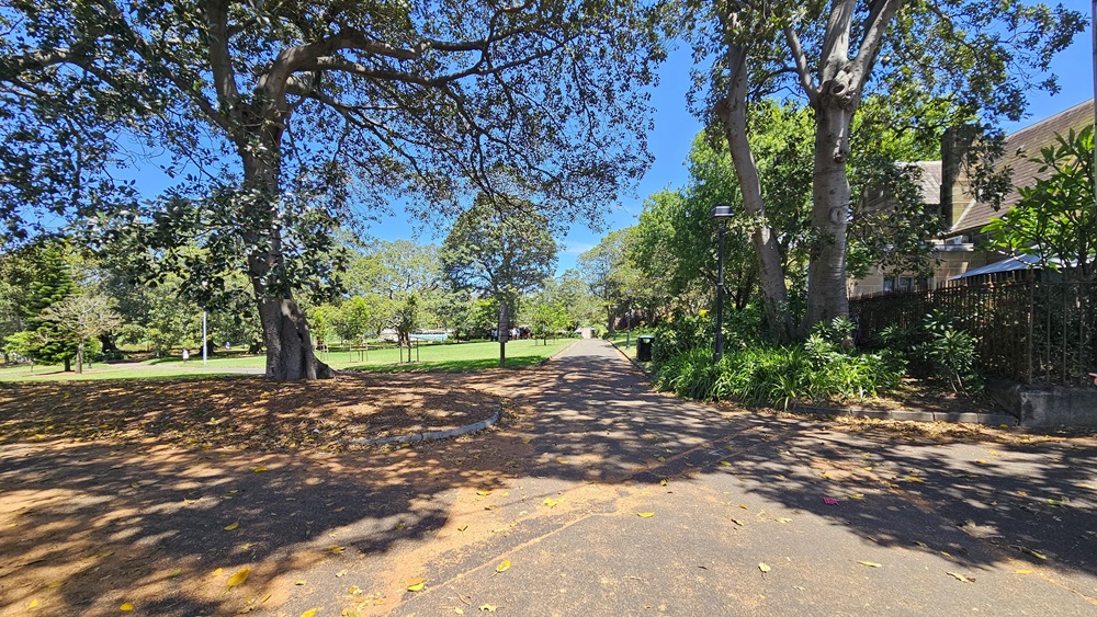 A straight brick path leads forward. To the right, there's a brick house enclosed by a metal fence. The right side is bordered by trees and shrubs. On the left, there's a view of green grass and tall trees. A straight brick path leads forward. To the right, there's a brick house enclosed by a metal fence. The right side is bordered by trees and shrubs. On the left, there's a view of green grass and tall trees.