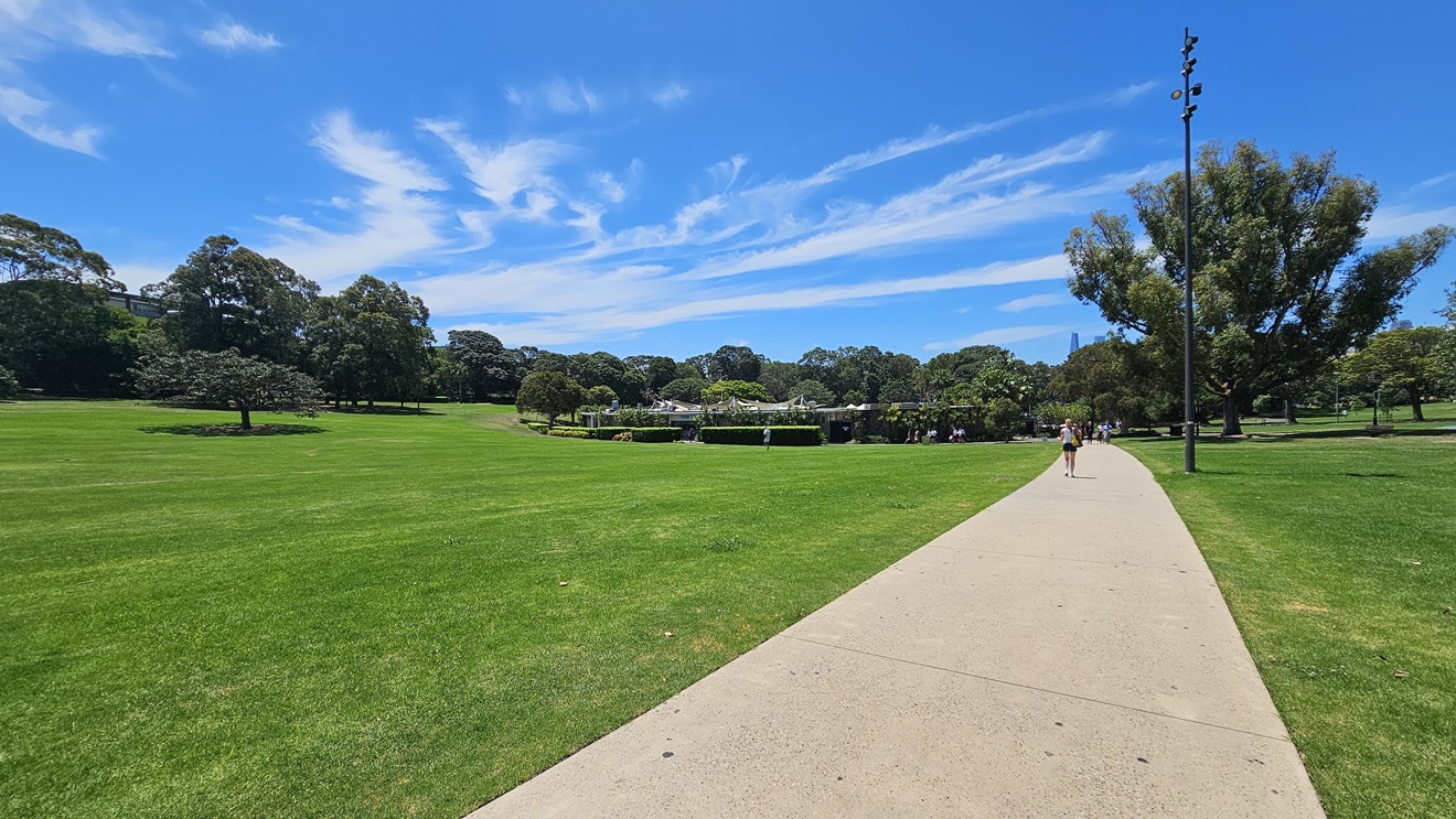 A straight concrete path is bordered by well-maintained grass and tall trees. A tall light post stands on the right. Beyond, there's a facility building. A person walking on the pathway is visible. A straight concrete path is bordered by well-maintained grass and tall trees. A tall light post stands on the right. Beyond, there's a facility building. A person walking on the pathway is visible.