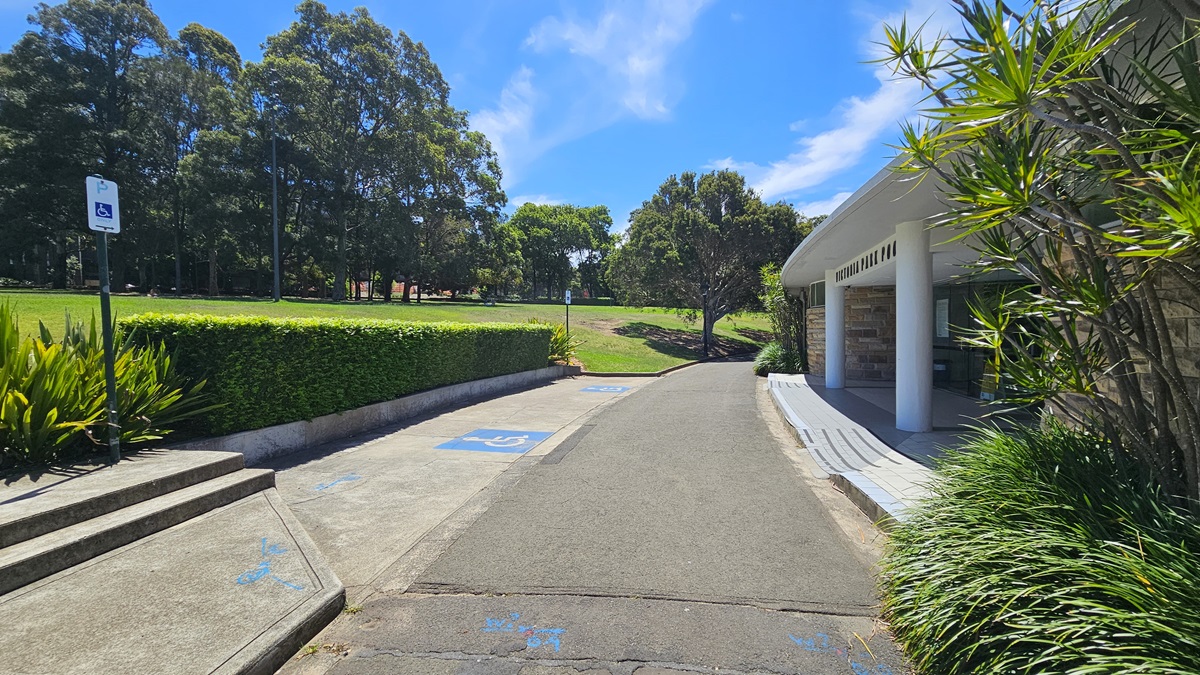 Accessible car parking spaces, marked by blue wheelchair signs, are located in front of the building. To the right, the modern brick structure is equipped with a step-free ramp. In front, there's a landscaped garden and neatly trimmed grass. An accessible sign is positioned on a raised platform near the landscaped shrubs. Tall trees and well-maintained grass surround the space. Accessible car parking spaces, marked by blue wheelchair signs, are located in front of the building. To the right, the modern brick structure is equipped with a step-free ramp. In front, there's a landscaped garden and neatly trimmed grass. An accessible sign is positioned on a raised platform near the landscaped shrubs. Tall trees and well-maintained grass surround the space.
