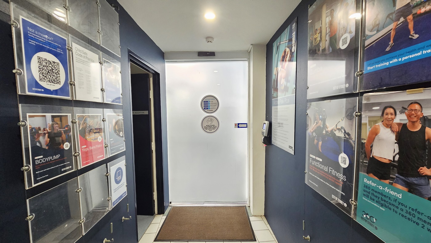The black wall is equipped with posters and directional signages. To the left is a white sliding door equipped with a swiping machine for badge entry. A brown mat is installed on the doorstep. To the right is a huge retractable banner. The floor is grey, and the ceiling and wall are white with lights. The black wall is equipped with posters and directional signages. To the left is a white sliding door equipped with a swiping machine for badge entry. A brown mat is installed on the doorstep. To the right is a huge retractable banner. The floor is grey, and the ceiling and wall are white with lights.