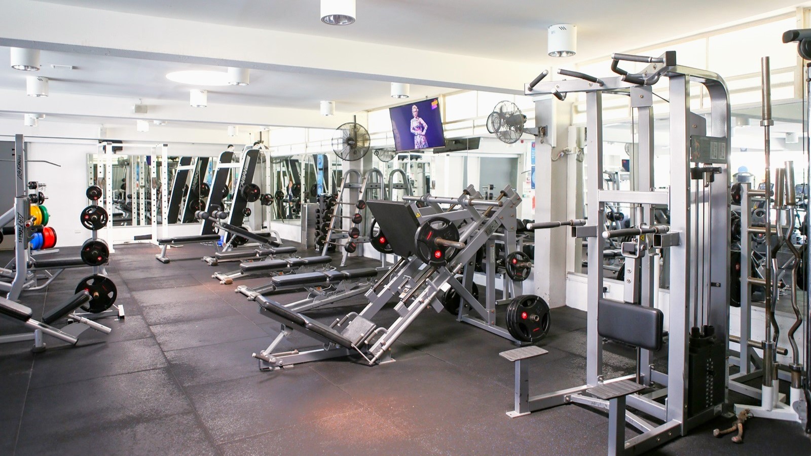Inside the gym, Lat Pulldown machines, Smith Machines, bench press, and inclined bench press machines are arranged on the black rubber-tiled floor. A flat-screen TV is mounted on the upper right wall along with wall fans. The ceiling is all white with multiple pin lights. Inside the gym, Lat Pulldown machines, Smith Machines, bench press, and inclined bench press machines are arranged on the black rubber-tiled floor. A flat-screen TV is mounted on the upper right wall along with wall fans. The ceiling is all white with multiple pin lights.