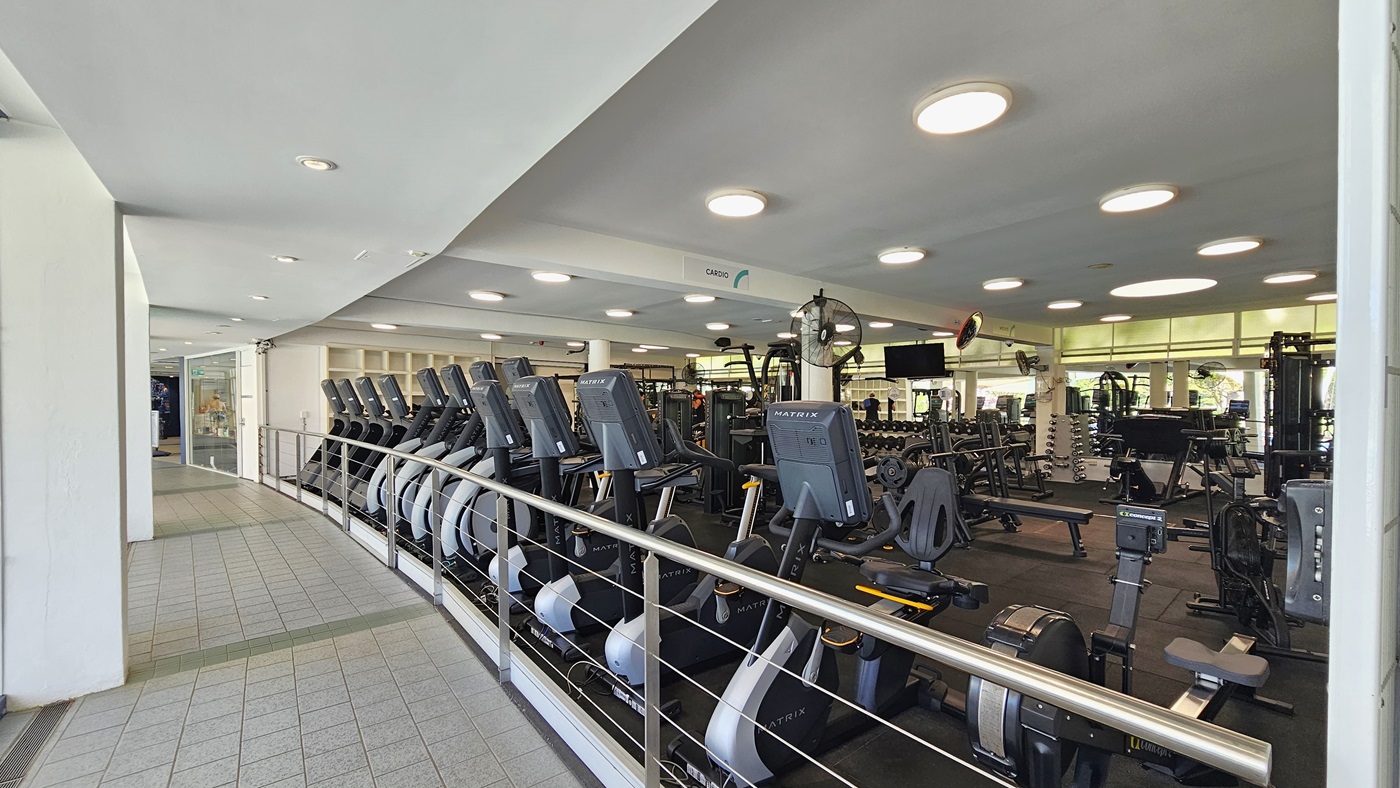 Cardio machines are arranged near the stainless railings. Inside, Lat Pulldown machines, Smith Machines, bench press, and inclined bench press machines, along with other machines, are arranged on the black rubber-tiled floor. A flat-screen TV and wall fans are affixed to the upper wall. To the left is a corridor with grey tiles. The ceiling is white with circular LED lights. Cardio machines are arranged near the stainless railings. Inside, Lat Pulldown machines, Smith Machines, bench press, and inclined bench press machines, along with other machines, are arranged on the black rubber-tiled floor. A flat-screen TV and wall fans are affixed to the upper wall. To the left is a corridor with grey tiles. The ceiling is white with circular LED lights.