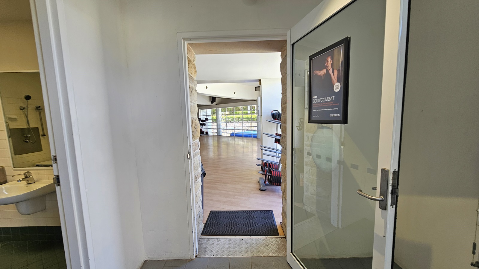 To the left is the bathroom, and adjacent to it is the Exercise Studio, accessible through a manual glass door equipped with a poster. An aluminum step-free ramp is installed on the doorstep along with a non-slippery mat. The studio features a timber floor, and inside, exercise machines are visible. To the left is the bathroom, and adjacent to it is the Exercise Studio, accessible through a manual glass door equipped with a poster. An aluminum step-free ramp is installed on the doorstep along with a non-slippery mat. The studio features a timber floor, and inside, exercise machines are visible.