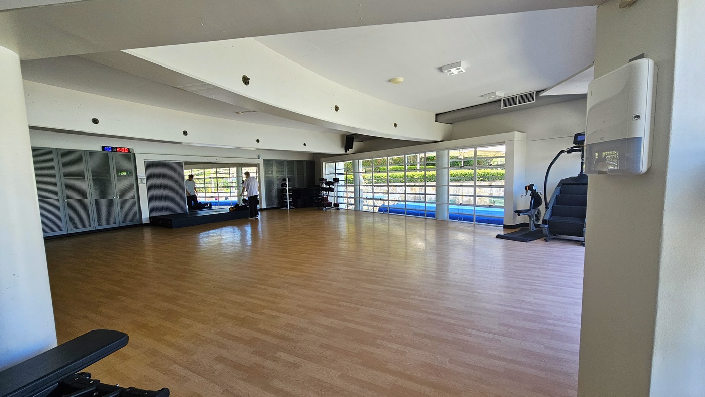 The exercise studio is a spacious area featuring a timber floor. Spin machines and a stair mill are on the right. Weights are arranged in the corner, and a platform is positioned against the mirror. A digital clock is affixed to the upper-left wall. The white ceiling features a cove design with LED lights. The exercise studio is a spacious area featuring a timber floor. Spin machines and a stair mill are on the right. Weights are arranged in the corner, and a platform is positioned against the mirror. A digital clock is affixed to the upper-left wall. The white ceiling features a cove design with LED lights.