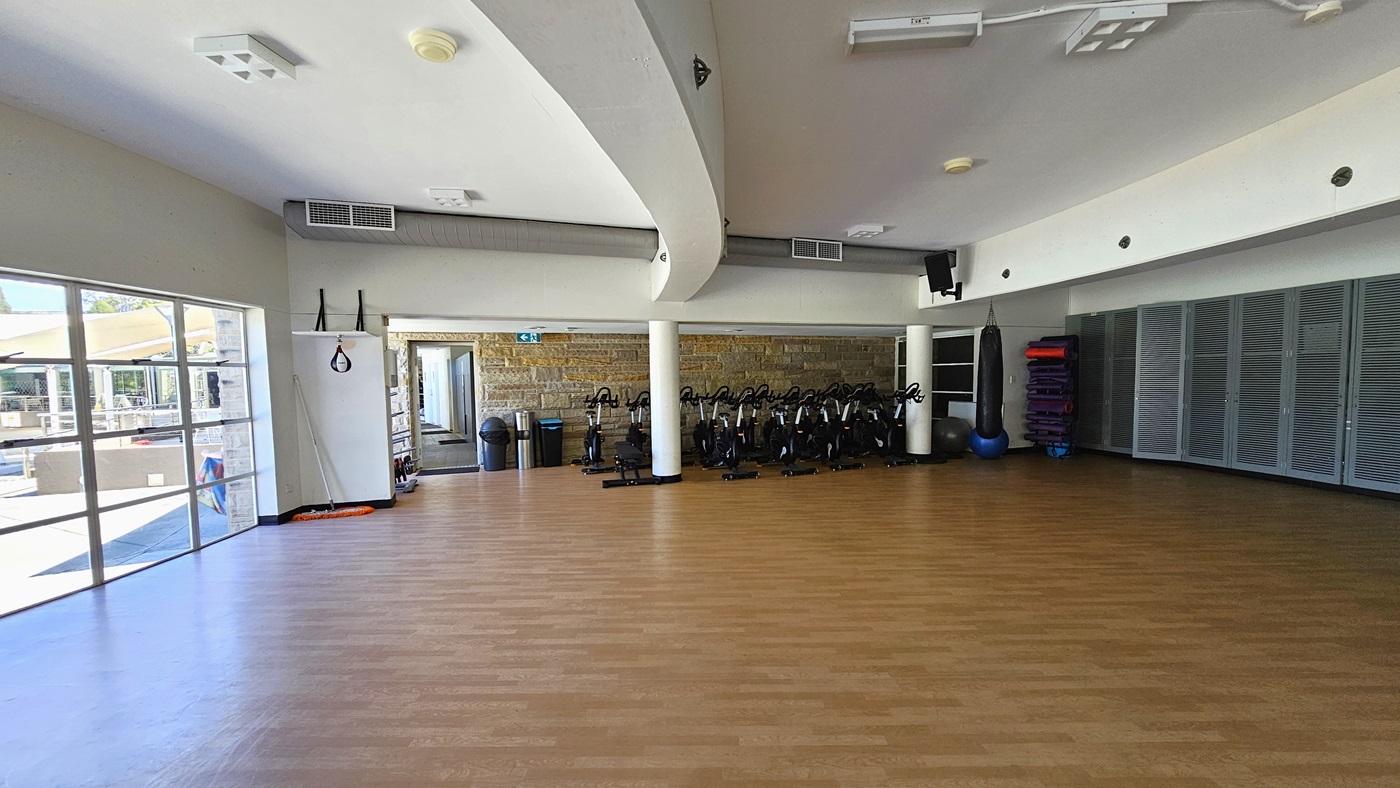 The exercise studio is a spacious area featuring a timber floor. The middle wall has brown bricks, and spin machines are arranged against it. Rubbish bins are placed near the door. In the right corner, there is a punching bag, rolled mats, and stability balls. The white ceiling features a cove design with LED lights. The left wall is made of glass. The exercise studio is a spacious area featuring a timber floor. The middle wall has brown bricks, and spin machines are arranged against it. Rubbish bins are placed near the door. In the right corner, there is a punching bag, rolled mats, and stability balls. The white ceiling features a cove design with LED lights. The left wall is made of glass.