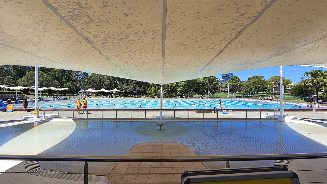 Access to the Outdoor Aquatic Facility is through a manual glass door. Immediately inside, free-standing banners and an aquatic wheelchair are positioned in the middle. Shade cloths overhead provide shade to the Kid's Splash Pool. An aluminum railing separates the upper pool deck and the pool area. Further behind is the lap pool, with pool guards and guests visible. Access to the Outdoor Aquatic Facility is through a manual glass door. Immediately inside, free-standing banners and an aquatic wheelchair are positioned in the middle. Shade cloths overhead provide shade to the Kid's Splash Pool. An aluminum railing separates the upper pool deck and the pool area. Further behind is the lap pool, with pool guards and guests visible.