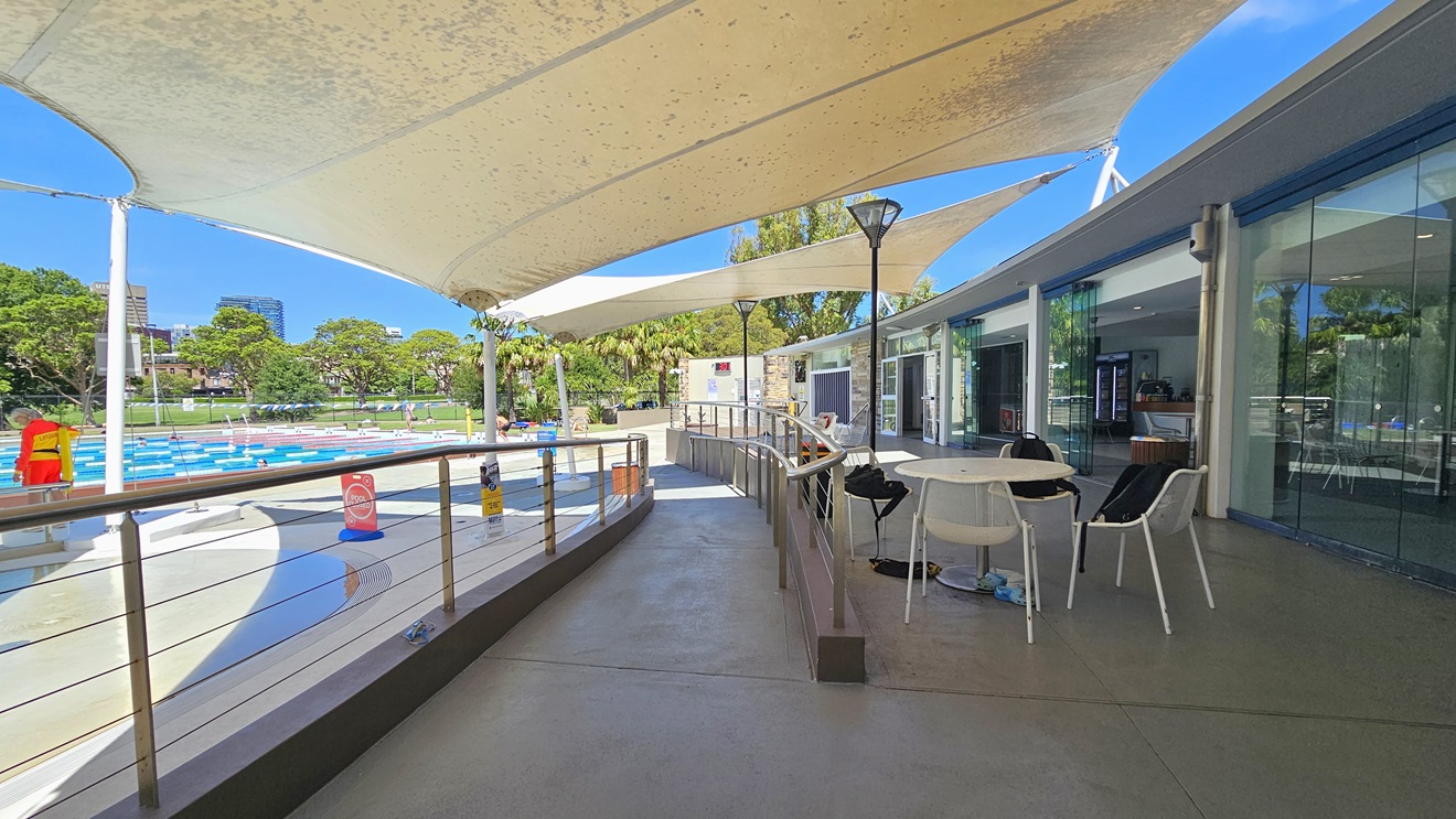 To the right is the cafe enclosed with glass. Outside, movable tables and chairs, equipped with belongings are displayed. A ramp entry, equipped with handrails, leads to the pool area. To the left are the Splash Pool and the 50m Lap Pool, equipped with floating ropes. White shade cloths are placed overhead, providing shade to the seating area. Pool guards and a free-standing banner are visible on the pool deck. Trees surround the area. To the right is the cafe enclosed with glass. Outside, movable tables and chairs, equipped with belongings are displayed. A ramp entry, equipped with handrails, leads to the pool area. To the left are the Splash Pool and the 50m Lap Pool, equipped with floating ropes. White shade cloths are placed overhead, providing shade to the seating area. Pool guards and a free-standing banner are visible on the pool deck. Trees surround the area.