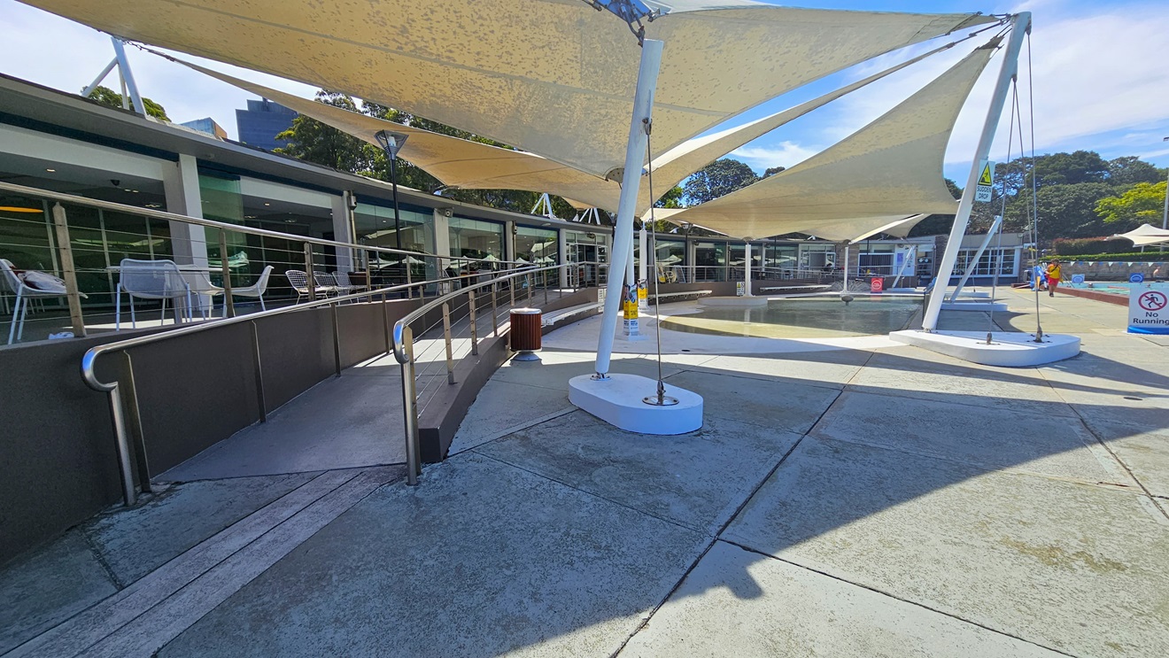 To the left is the cafe enclosed with glass. Outside, movable tables and chairs are displayed. A ramp entry, equipped with handrails, leads to the pool area. To the right are the Splash Pool and the 50m Lap Pool, equipped with floating ropes. White shade cloths are placed overhead, providing shade to the seating area. Pool guards and a free-standing banner are visible on the pool deck. Trees surround the area. To the left is the cafe enclosed with glass. Outside, movable tables and chairs are displayed. A ramp entry, equipped with handrails, leads to the pool area. To the right are the Splash Pool and the 50m Lap Pool, equipped with floating ropes. White shade cloths are placed overhead, providing shade to the seating area. Pool guards and a free-standing banner are visible on the pool deck. Trees surround the area.