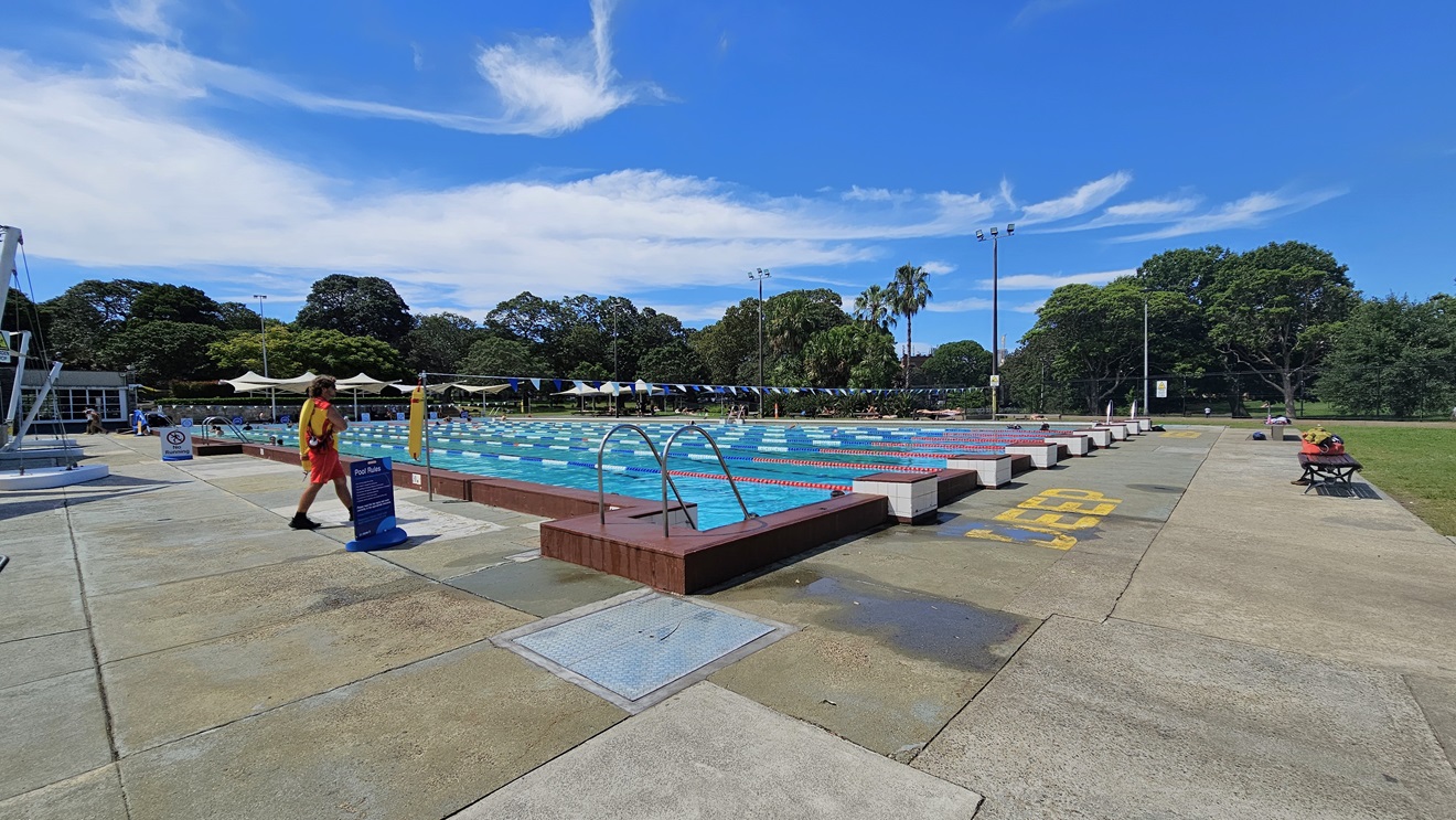 The 50m Lap Pool is equipped with floating ropes and step entry points with handrails. Bench seating with belongings is placed on the right near the grass. The concrete floor has yellow markings indicating the pool's deep water level. Free-standing banners and safety warnings are on the pool deck. A string of blue buntings runs across the pool. Further ahead, umbrella shades are visible, and a pool staff member is present. The 50m Lap Pool is equipped with floating ropes and step entry points with handrails. Bench seating with belongings is placed on the right near the grass. The concrete floor has yellow markings indicating the pool's deep water level. Free-standing banners and safety warnings are on the pool deck. A string of blue buntings runs across the pool. Further ahead, umbrella shades are visible, and a pool staff member is present.