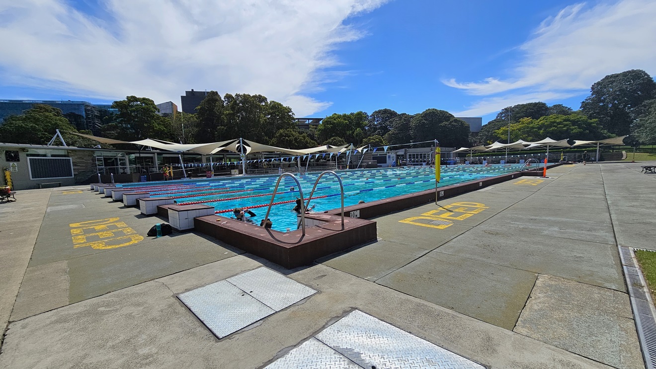 The 50m Lap Pool features floating ropes and step entry points with handrails. The concrete floor is marked with yellow indications for the deep water level. Free-standing banners and safety warnings are present on the pool deck. A string of blue buntings runs across the pool. Further ahead, umbrella shades are visible on the left and far right. The 50m Lap Pool features floating ropes and step entry points with handrails. The concrete floor is marked with yellow indications for the deep water level. Free-standing banners and safety warnings are present on the pool deck. A string of blue buntings runs across the pool. Further ahead, umbrella shades are visible on the left and far right.