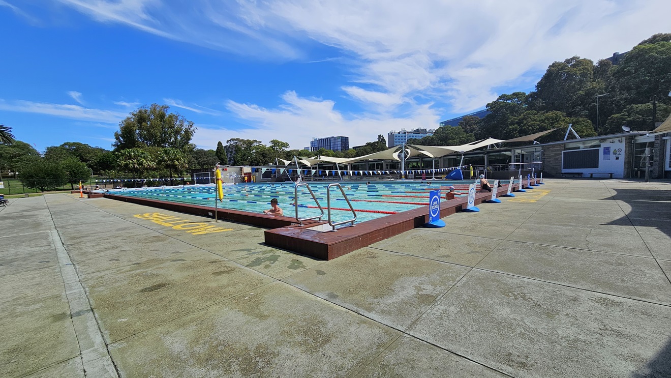 The 50m Lap Pool is equipped with floating ropes and step entry points with handrails. The concrete floor is marked with yellow indications for the shallow water level. Free-standing banners are positioned on the pool deck. A string of blue buntings runs across the pool. Further ahead, umbrella shades are visible on the right, adjacent to a brick building. Tall trees surround the area. The 50m Lap Pool is equipped with floating ropes and step entry points with handrails. The concrete floor is marked with yellow indications for the shallow water level. Free-standing banners are positioned on the pool deck. A string of blue buntings runs across the pool. Further ahead, umbrella shades are visible on the right, adjacent to a brick building. Tall trees surround the area.