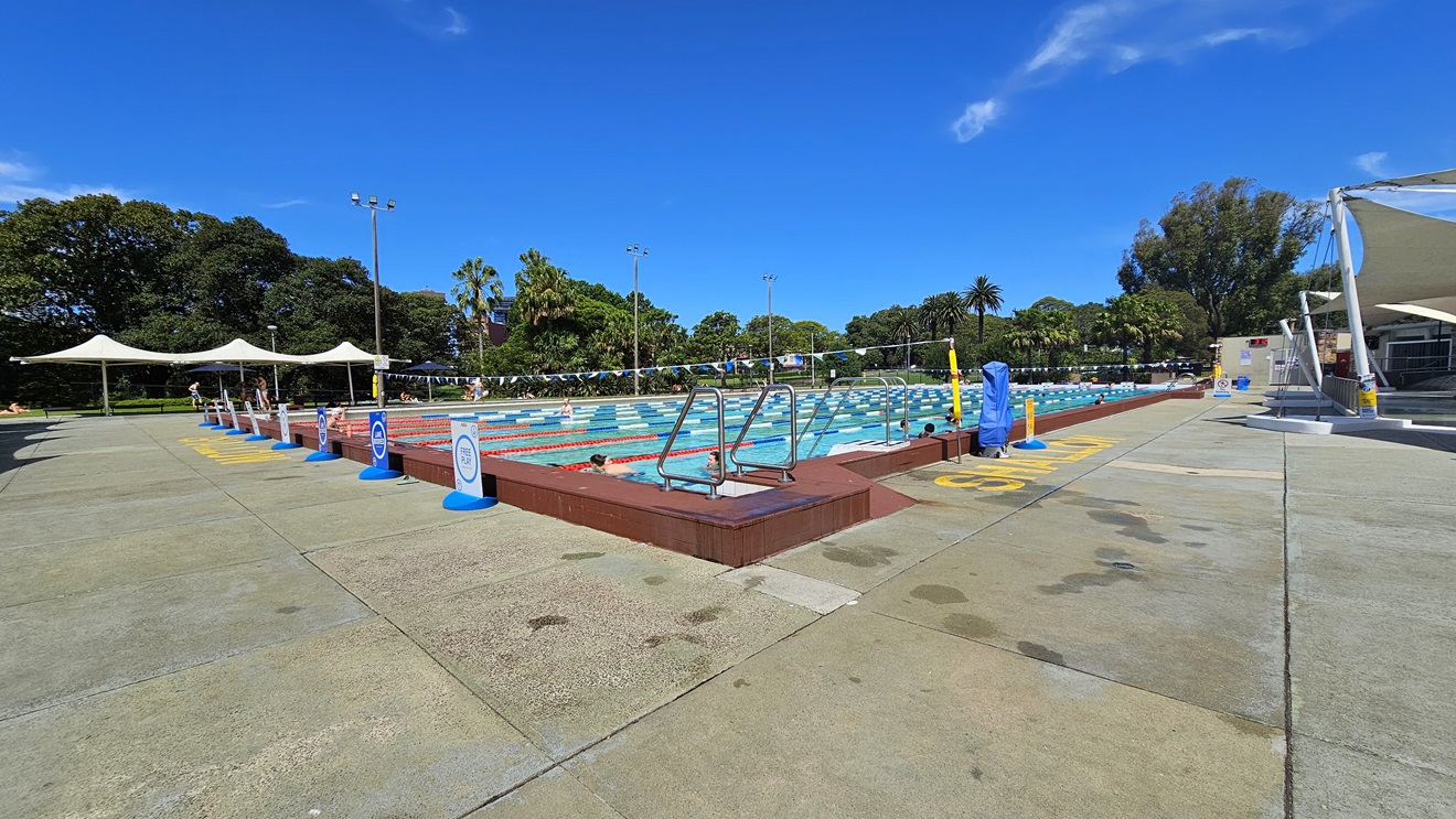 The 50m Lap Pool has floating ropes and step entry points with handrails. The concrete floor is marked with yellow indications for the shallow water level. On the left, free-standing banners are positioned on the pool deck. A string of blue buntings runs across the pool. To the left, umbrella shades are visible, and shade cloths are on the right. A hoist is positioned near the pool. Tall trees surround the area. The 50m Lap Pool has floating ropes and step entry points with handrails. The concrete floor is marked with yellow indications for the shallow water level. On the left, free-standing banners are positioned on the pool deck. A string of blue buntings runs across the pool. To the left, umbrella shades are visible, and shade cloths are on the right. A hoist is positioned near the pool. Tall trees surround the area.