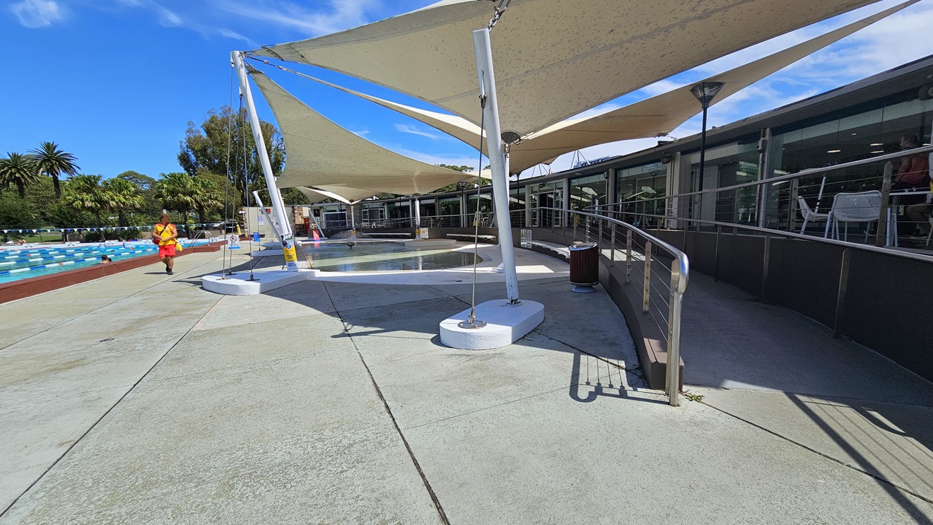 To the right is the cafe enclosed with glass. Outside, movable tables and chairs are displayed. A ramp entry, equipped with handrails, leads to the pool area. To the left is the 50m Lap Pool, equipped with floating ropes. White shade cloths are placed overhead, providing shade to the seating area. Pool guards and a free-standing banner are visible on the pool deck. Trees surround the area. To the right is the cafe enclosed with glass. Outside, movable tables and chairs are displayed. A ramp entry, equipped with handrails, leads to the pool area. To the left is the 50m Lap Pool, equipped with floating ropes. White shade cloths are placed overhead, providing shade to the seating area. Pool guards and a free-standing banner are visible on the pool deck. Trees surround the area.