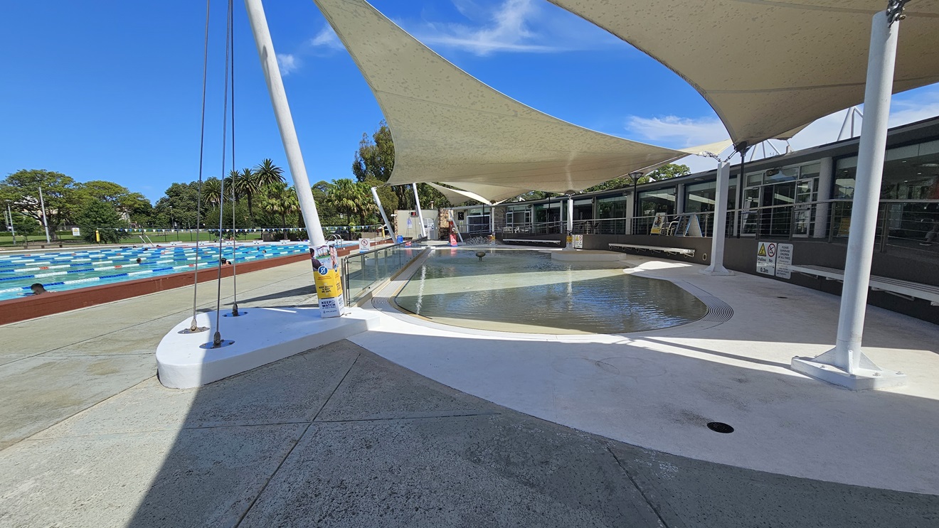 To the right is a ramp entry, equipped with handrails, leading to the pool area. In the middle is the Kid's Splash Pool with a beach entry and a small water fountain. To the left is the 50m Lap Pool, equipped with floating ropes. White shade cloths are placed overhead, providing shade to the seating area. Free-standing banners and safety signs are visible. Trees surround the area. To the right is a ramp entry, equipped with handrails, leading to the pool area. In the middle is the Kid's Splash Pool with a beach entry and a small water fountain. To the left is the 50m Lap Pool, equipped with floating ropes. White shade cloths are placed overhead, providing shade to the seating area. Free-standing banners and safety signs are visible. Trees surround the area.