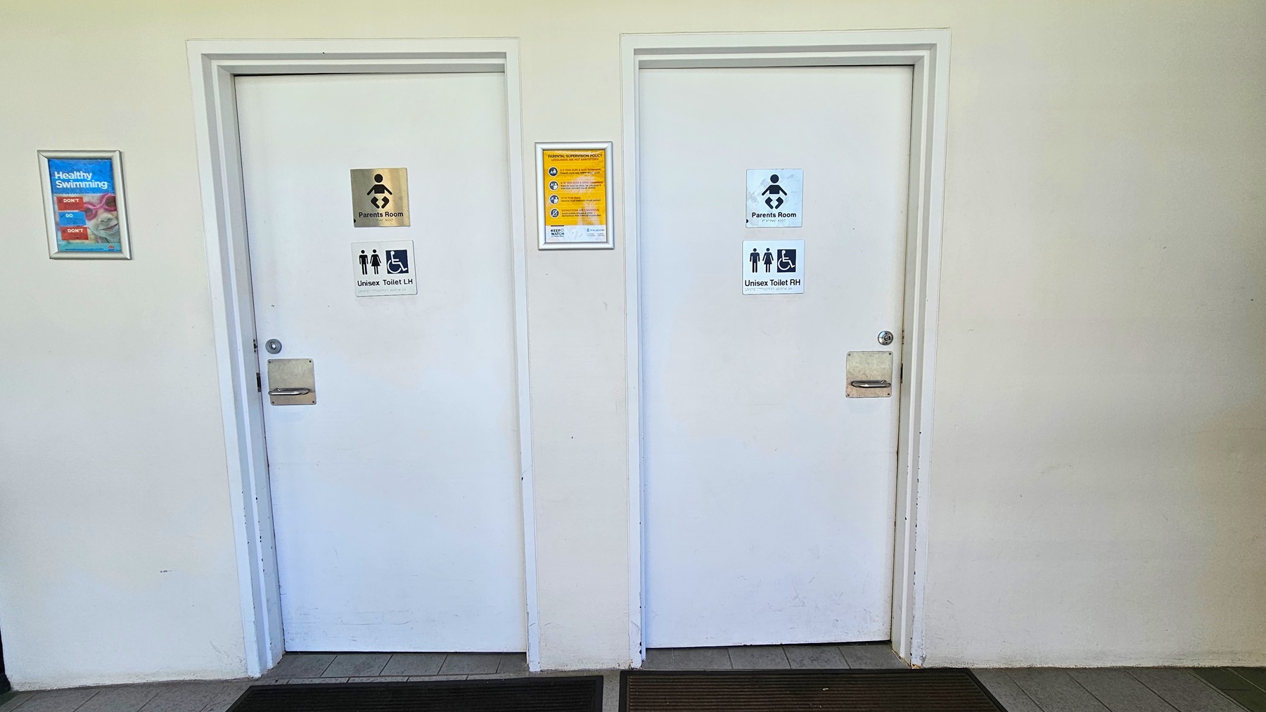 wo white doors lead to the accessible unisex bathroom and change rooms. A poster is affixed to the middle and to the right. Non-slippery mats are placed on the doorsteps. The wall is white, and the floor has grey tiles. wo white doors lead to the accessible unisex bathroom and change rooms. A poster is affixed to the middle and to the right. Non-slippery mats are placed on the doorsteps. The wall is white, and the floor has grey tiles.