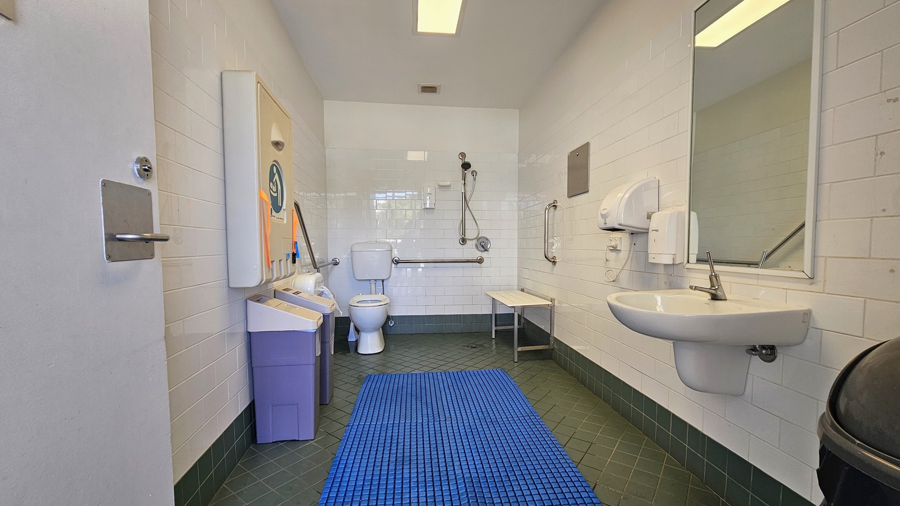 Inside the accessible bathroom, the toilet bowl equipped with handrails is in the left corner. Adjacent to it is the shower, with hand rails and a foldable bench affixed to the middle wall. To the right is a washbasin, mirrors, along with a liquid soap and tissue dispenser. A foldable change table is mounted to the left wall, and below it are two rubbish bins. The grey floor is equipped with a blue non-slippery mat, and the ceiling and wall are white. Inside the accessible bathroom, the toilet bowl equipped with handrails is in the left corner. Adjacent to it is the shower, with hand rails and a foldable bench affixed to the middle wall. To the right is a washbasin, mirrors, along with a liquid soap and tissue dispenser. A foldable change table is mounted to the left wall, and below it are two rubbish bins. The grey floor is equipped with a blue non-slippery mat, and the ceiling and wall are white.
