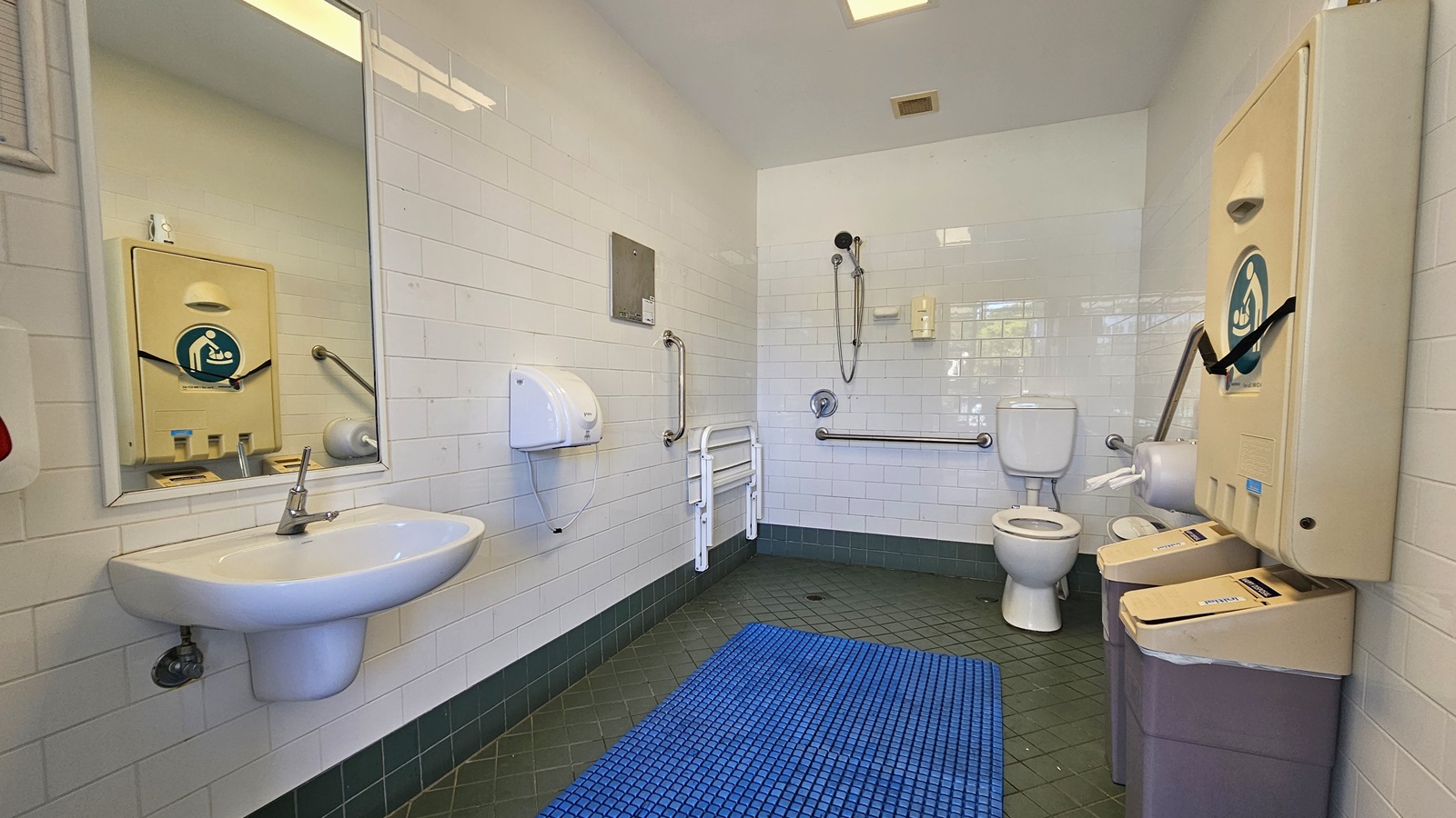 Inside the accessible bathroom, the toilet bowl equipped with handrails is in the right corner. Adjacent to it is the shower, with hand rails and a foldable bench affixed to the left wall. To the left is a washbasin, mirrors, along with a liquid soap and tissue dispenser. A foldable change table is mounted to the right wall, and below it are two rubbish bins. The grey floor is equipped with a blue non-slippery mat, and the ceiling and wall are white. Inside the accessible bathroom, the toilet bowl equipped with handrails is in the right corner. Adjacent to it is the shower, with hand rails and a foldable bench affixed to the left wall. To the left is a washbasin, mirrors, along with a liquid soap and tissue dispenser. A foldable change table is mounted to the right wall, and below it are two rubbish bins. The grey floor is equipped with a blue non-slippery mat, and the ceiling and wall are white.